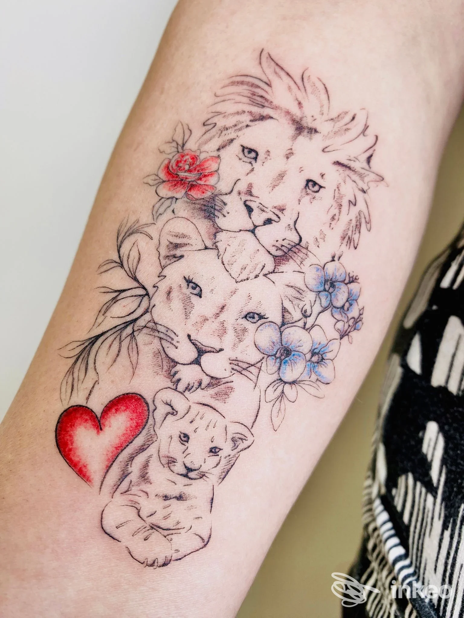 Tatuaggio su avambraccio con tre teste di leone, fiori blu e rosa e un cuore rosso, opera presentata da Giovanna Bacchin, Udine.