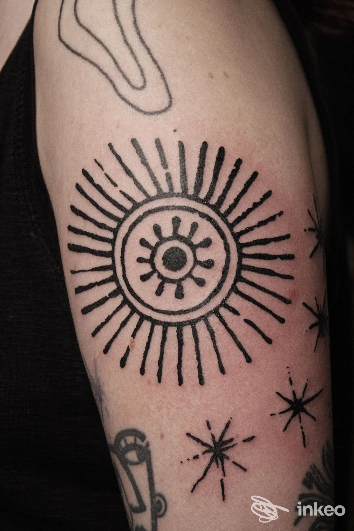 Tatuaggio a inchiostro nero su braccio: grande sole stilizzato con raggi e piccole stelle. New Nazareth Tattoo Studio, Udine.