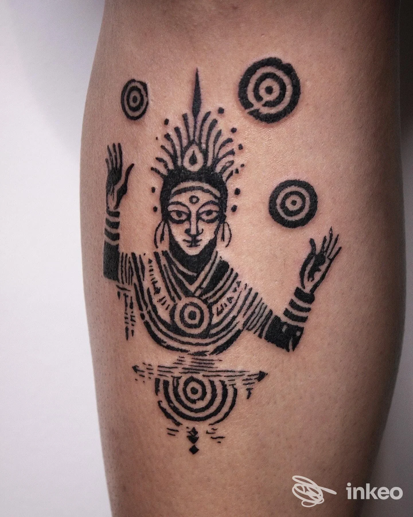 Tatuaggio nero stilizzato di figura divina con simboli circolari sul polpaccio. New Nazareth Tattoo Studio, Udine.