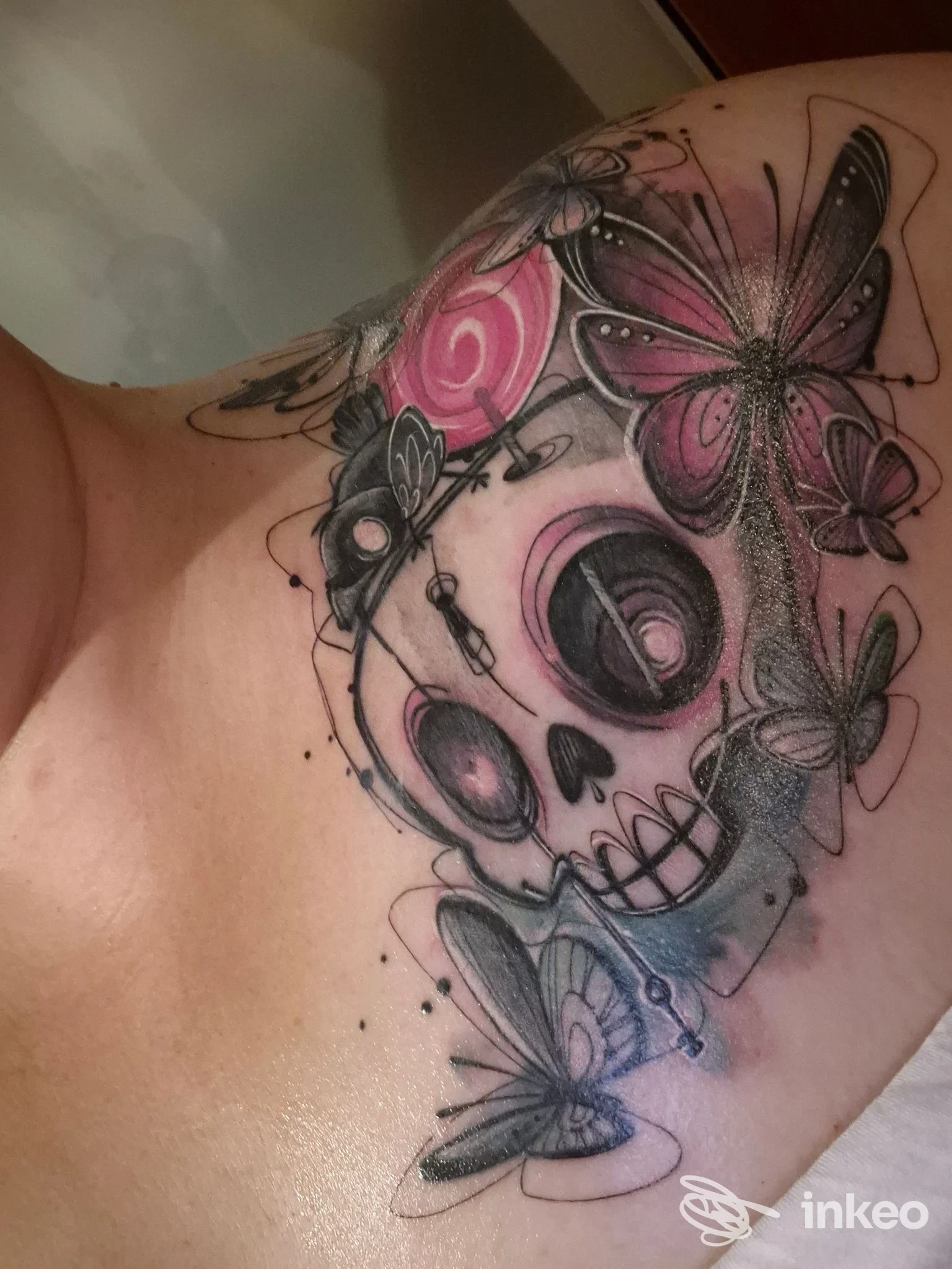 Tatuaggio sul petto con teschio stilizzato, farfalle e fiori in nero, grigio e rosa – Amentattoo, Udine.