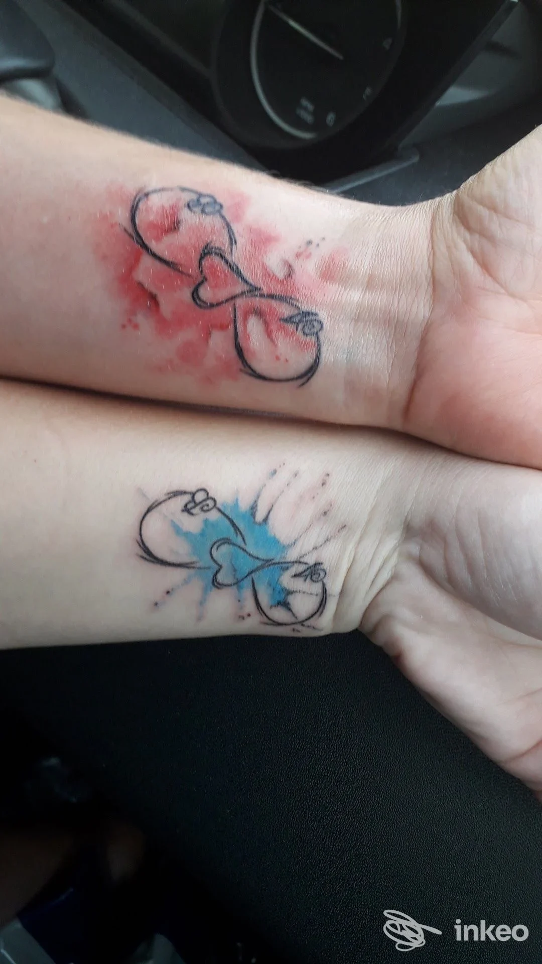 Due piccoli tatuaggi a forma di cuore con contorni neri e macchie acquerellate rosse e blu sui polsi, foto fornita da Pain Hill Tattoo a Montereale Valcellina.