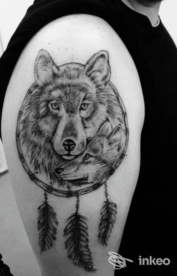 Tatuaggio realistico in bianco e nero su braccio superiore: due lupi in un acchiappasogni con piume. Jolie Tattoo Piercing Estetica, Udine.