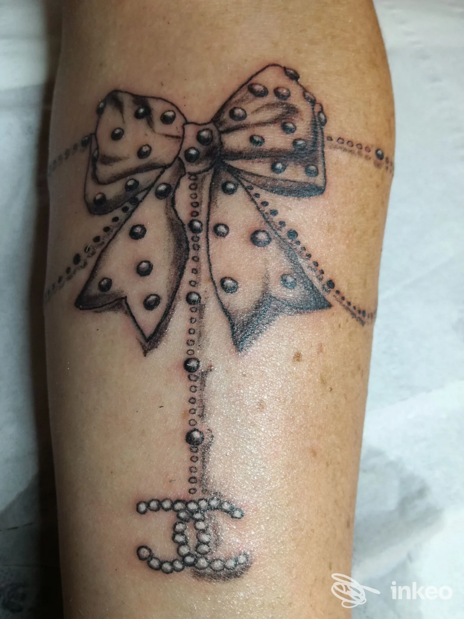 Tatuaggio su avambraccio: fiocco a pois con catenina di perle e monogramma, Sunflower Tattoo, Basiliano.