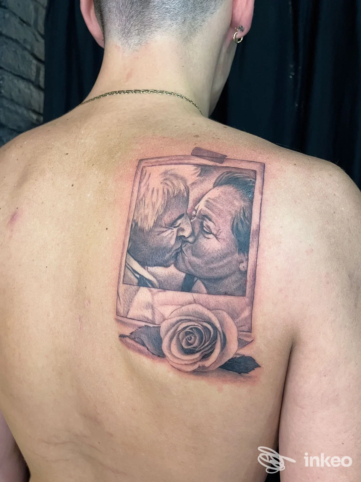Tattoo sul dorso raffigurante una cornice con due persone che si baciano e una rosa in basso, eseguito in tonalità nere/grigie.