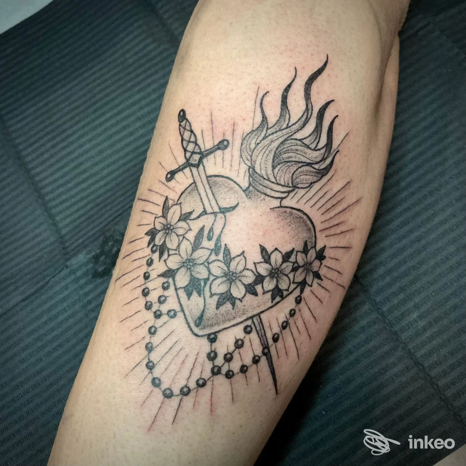 Tatuaggio a inchiostro nero di cuore fiammeggiante trafitto con fiori e rosario sull'avambraccio. Obsidian Tattoo Atelier, Trieste.