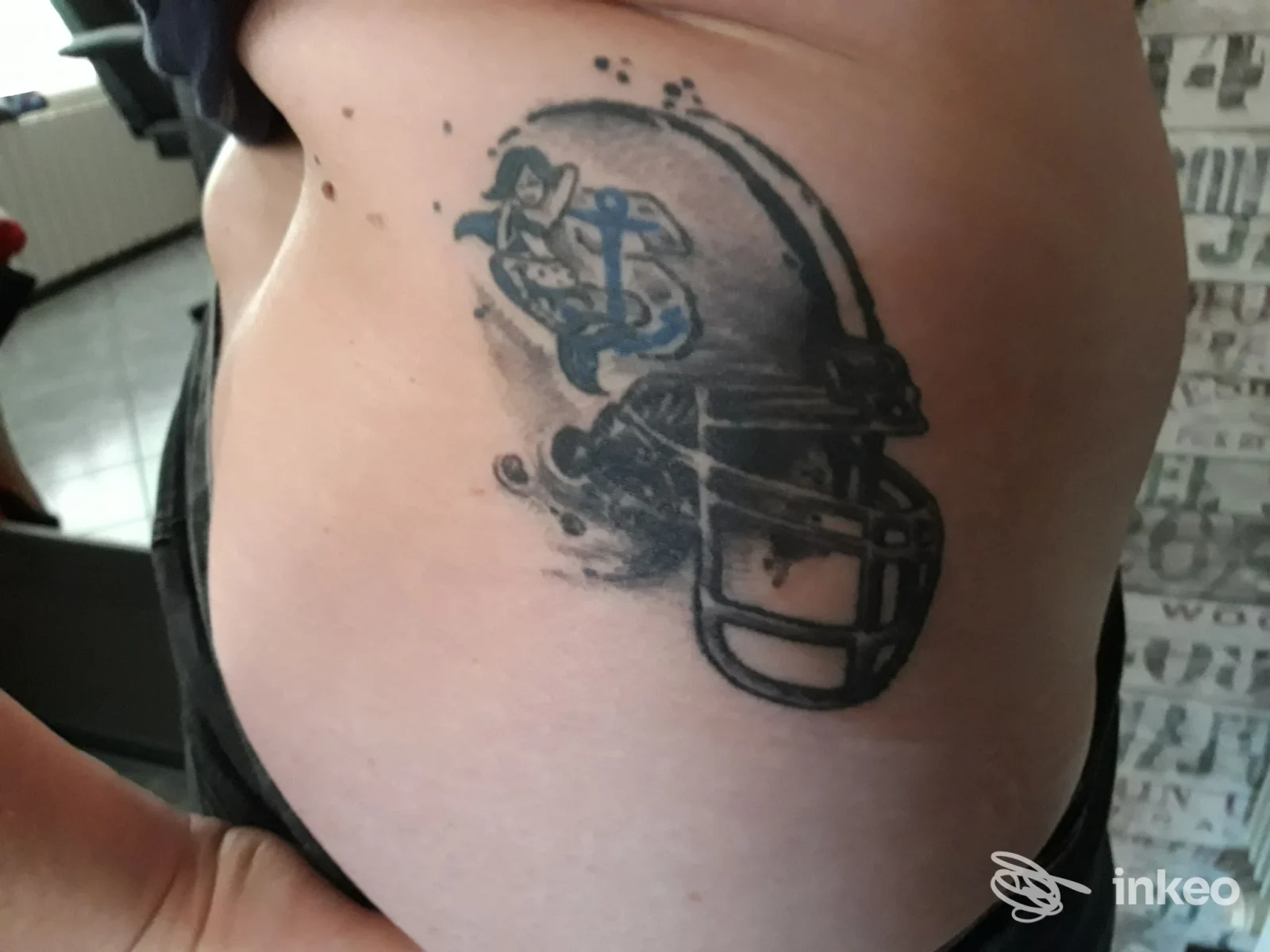 Tattoo su pelle visibile sul fianco raffigurante un casco da football accompagnato da una barra decorativa, realizzato in tonalità scure; nessun elemento di social media presente.