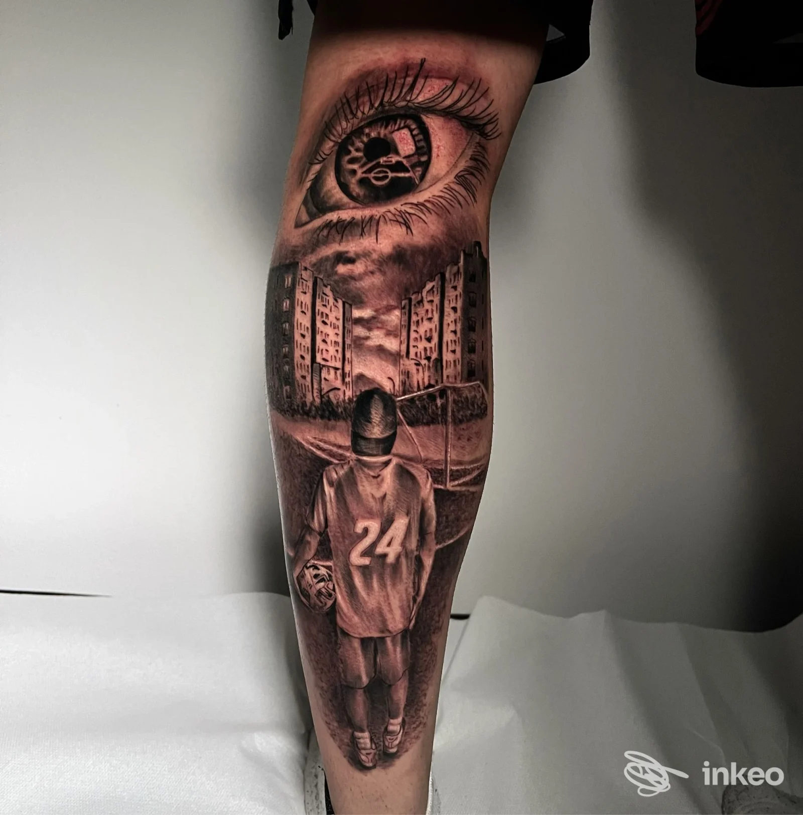 Tatuaggio realistico in bianco e nero su polpaccio con occhio meccanico, palazzi e figura con maglia numero 24 — Pure Body Art, Trieste.