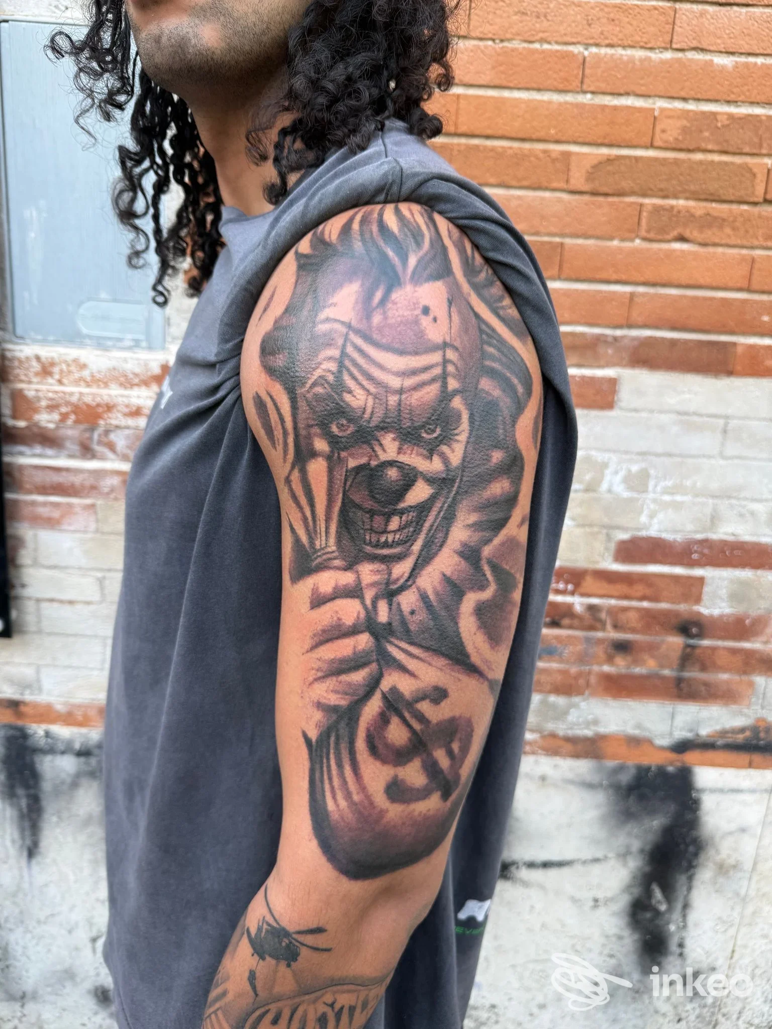 Primo piano del braccio con grande tatuaggio clown inquietante su un uomo, fotografato davanti a un muro di mattoni. Carlos'ink tattoo shop, Cassino.