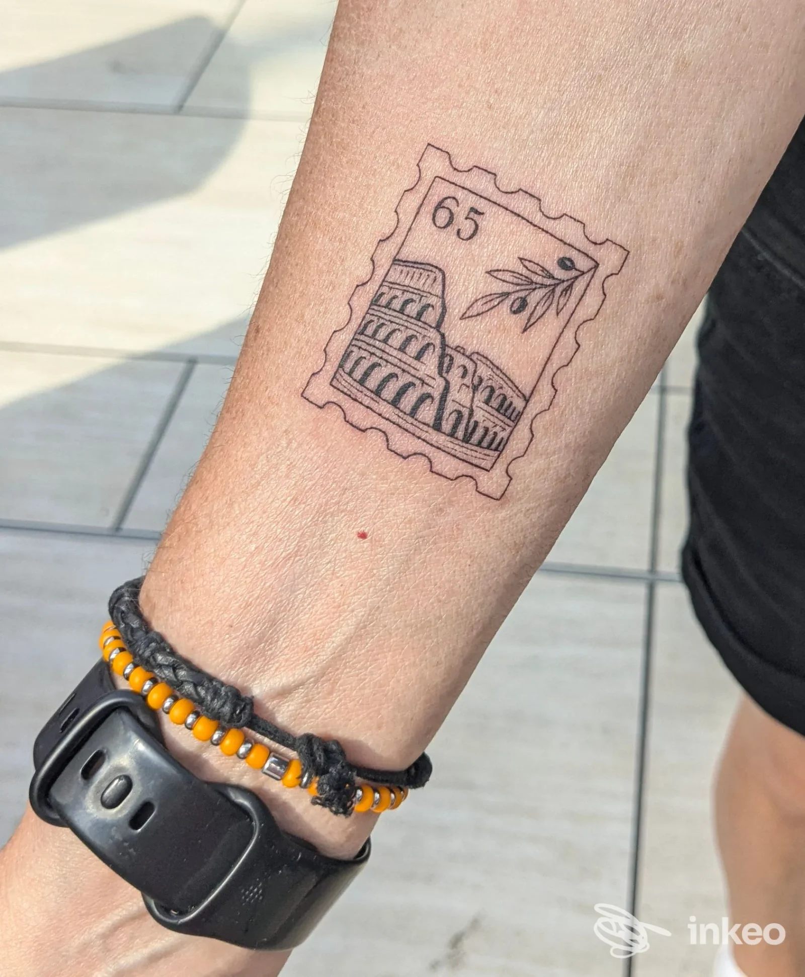 Tatuaggio a linee sottili a forma di francobollo con il Colosseo e ramo d'ulivo sull'avambraccio, foto inviata da Domus Tattoo, Roma.