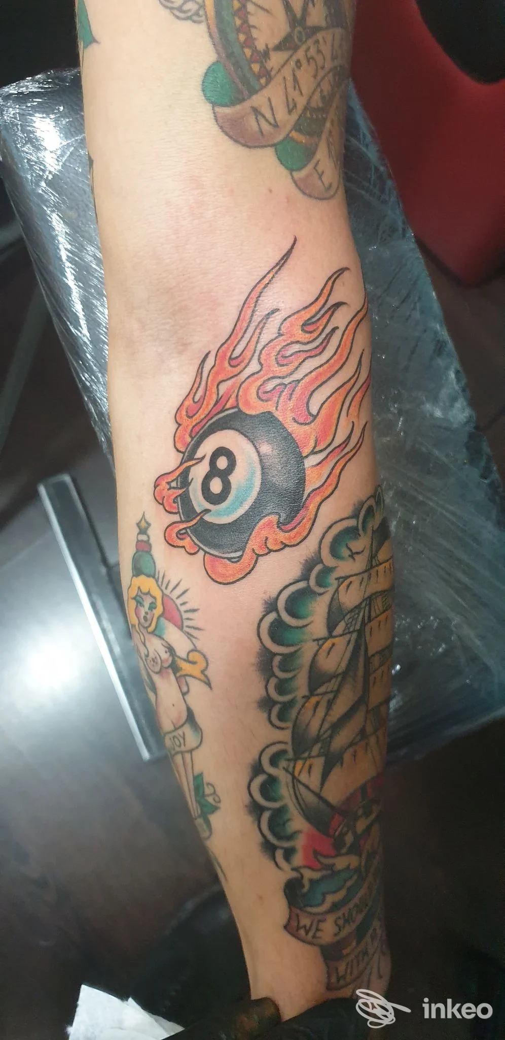 Avambraccio con diversi tatuaggi in stile tradizionale; al centro palla da biliardo n.8 con fiamme. Immagine per Fu Manchu Tattoo Palestrina, Palestrina.