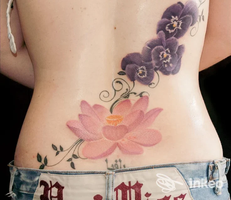 Tatuaggio floreale sul basso della schiena con loto rosa e fiori viola, foto fornita da Moko Tattoo Studio, Roma.