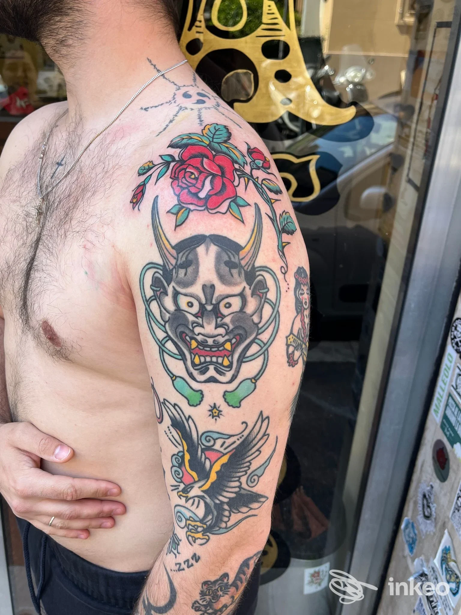 Spalla e braccio tatuati con rosa, maschera demoniaca e rondine, foto scattata davanti alla vetrina di Old Boat Tattoo a Roma.