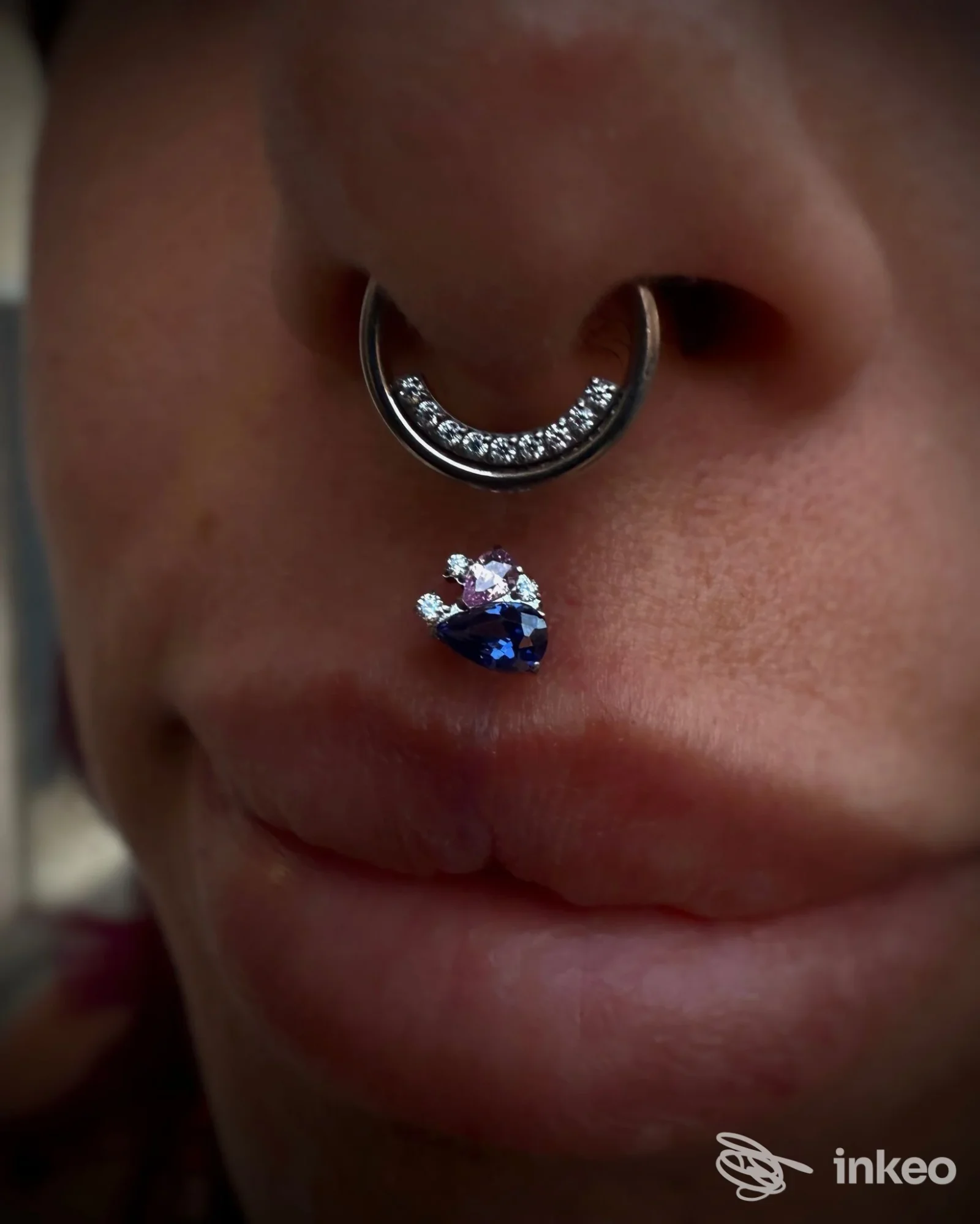 Primo piano di un setto nasale e philtrum con piercing (Perla bodypiercing & jewelry, Roma). Anello decorato e gioiello a goccia visibili sulla pelle.
