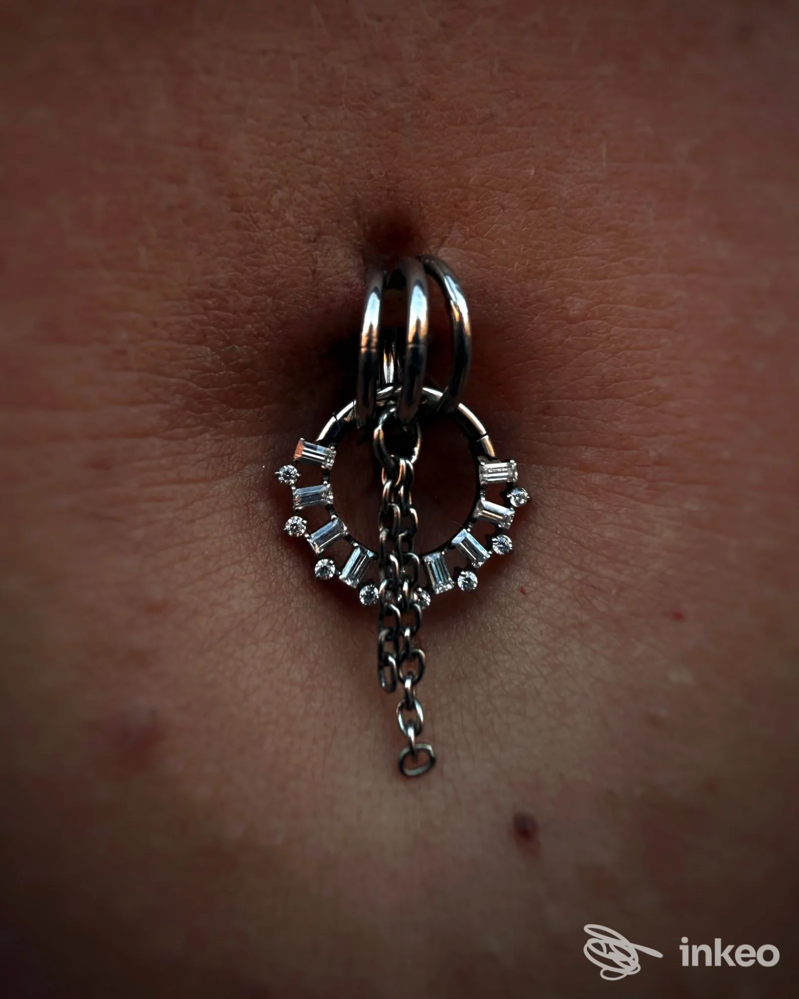 Piercing ombelicale con gioiello a cerchio decorato e catenella, fotografato su pelle; immagine da Perla bodypiercing & jewelry, Roma.