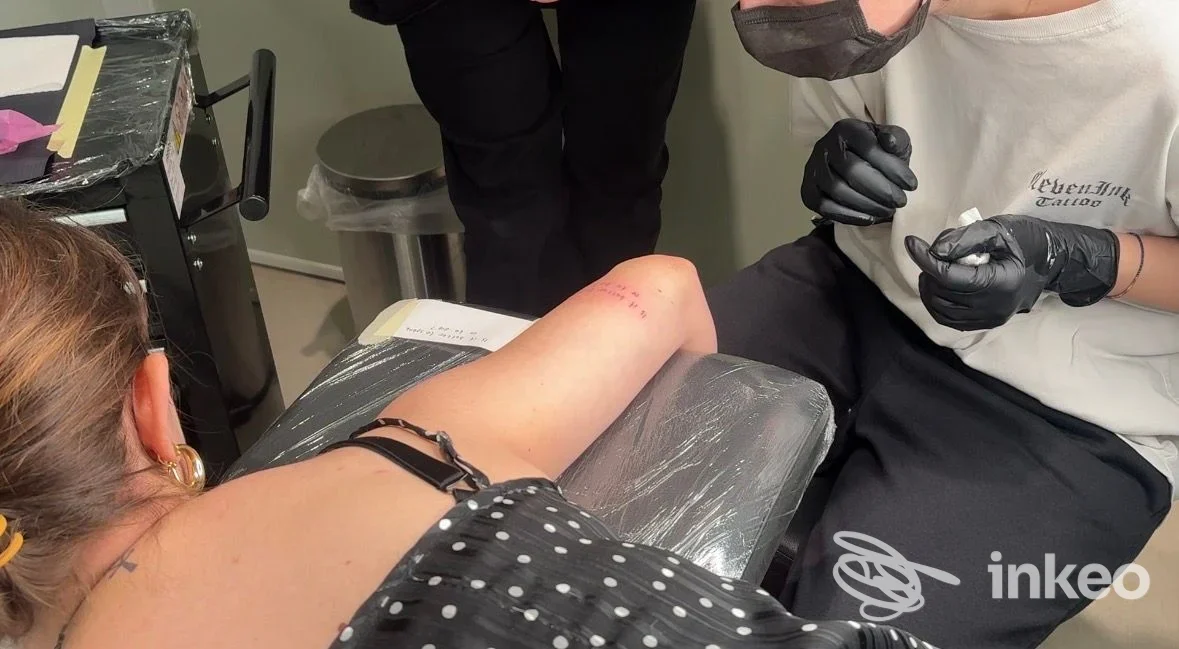 Un tatuaggio è in corso sull'avambraccio di una persona seduta; l'artista indossa guanti neri e lavora con la macchina per tatuare, in un ambiente di studio.