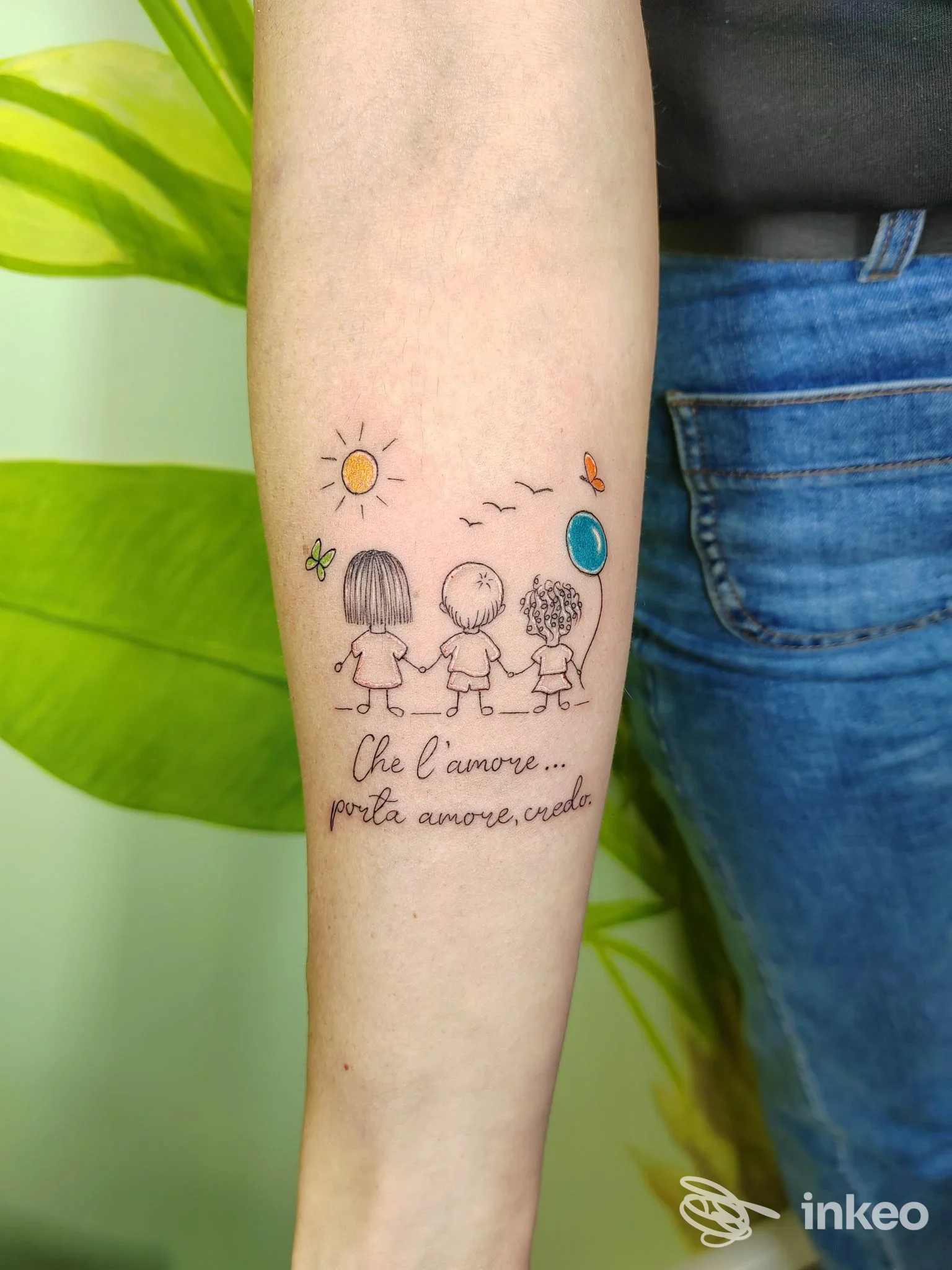Primo piano di un tatuaggio sull'avambraccio con tre bambini stilizzati, piccoli dettagli colorati e la scritta “Che l'amore... porta amore, crea”, foto per Tattooeasy3 a Roma.