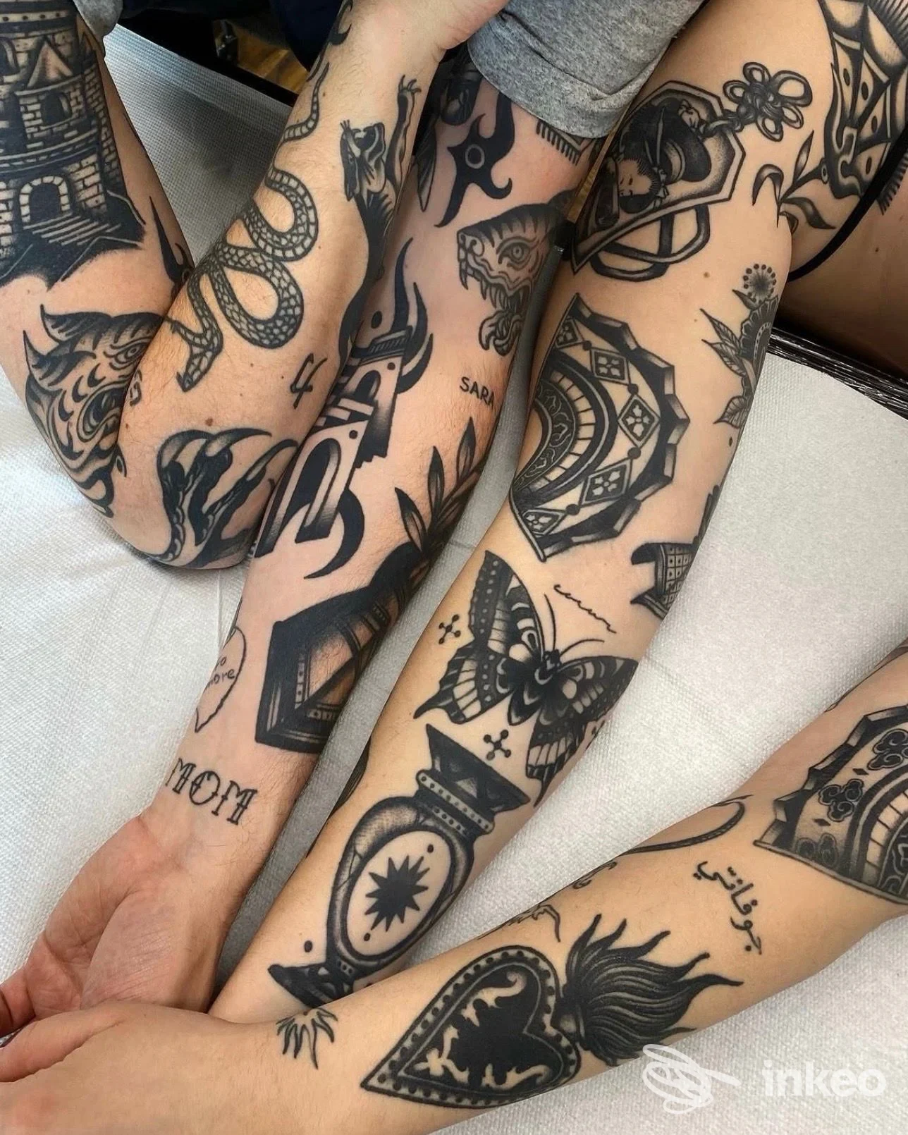 Braccia con vari tatuaggi in stile linework, tra cui uccello, farfalla e architetture, visibili su avambracci intrecciati, in primo piano.