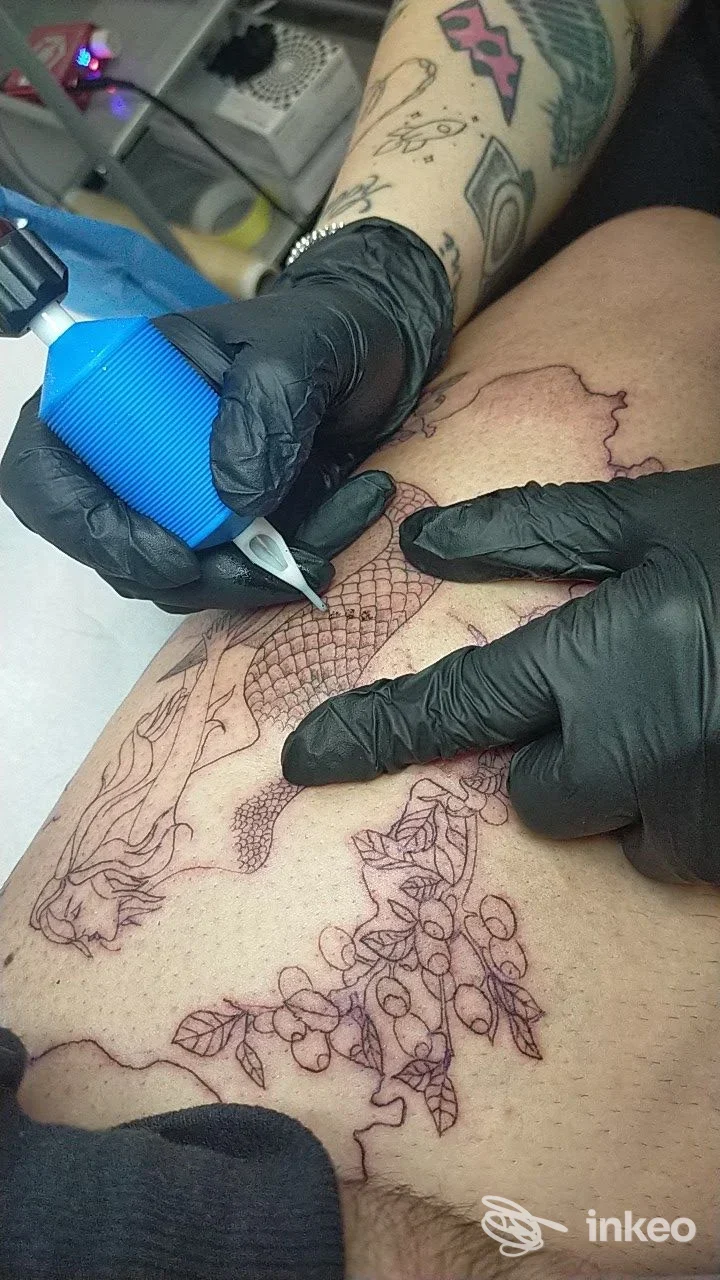 Primo piano di una seduta di tatuaggio sulla coscia, mani con guanti neri e macchinetta blu; Violet tattoo studio, Roma.