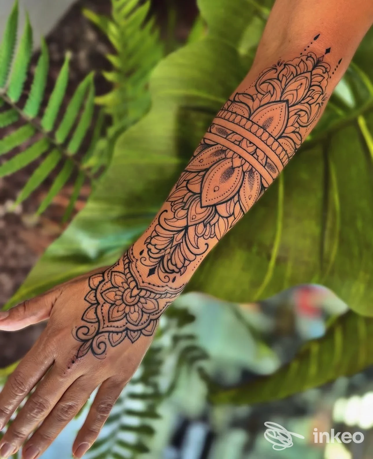 Tattoo elaborato a mandala sull’avambraccio sinistro, disegno nero su pelle, senza volto visibile, sfondo di foglie verdi.