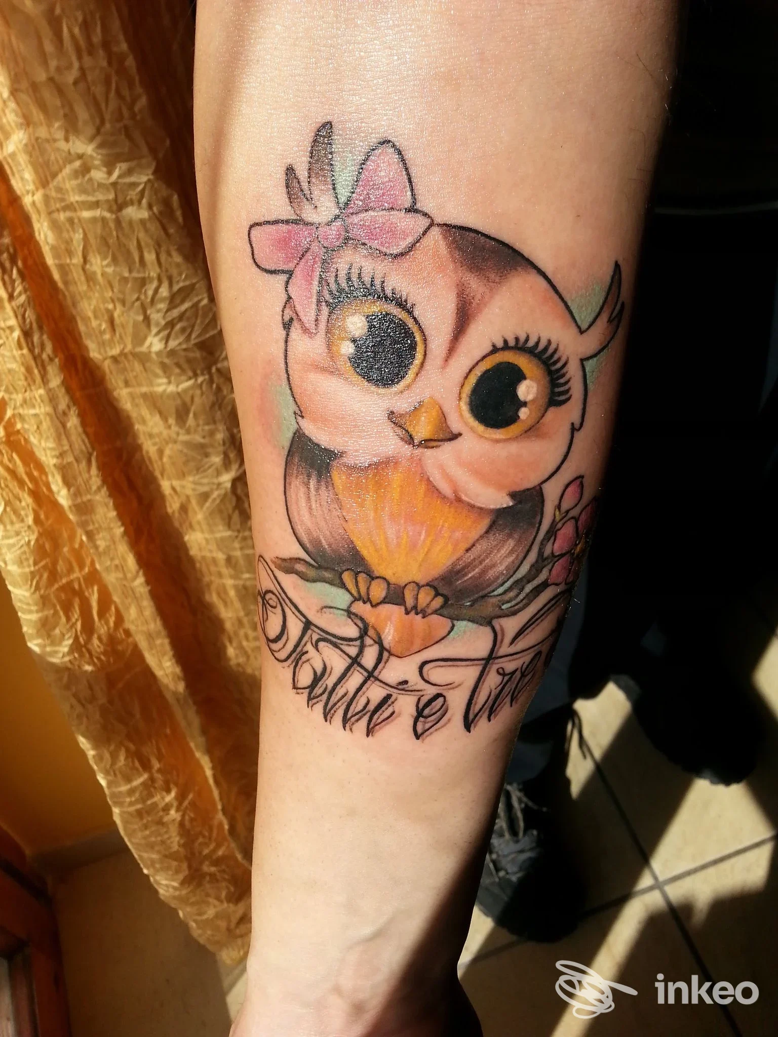 Tatuaggio sul avambraccio di un gufetto colorato con fiocco e fiori, foto fornita da 8th SIN TATTOO, Lido di Ostia.