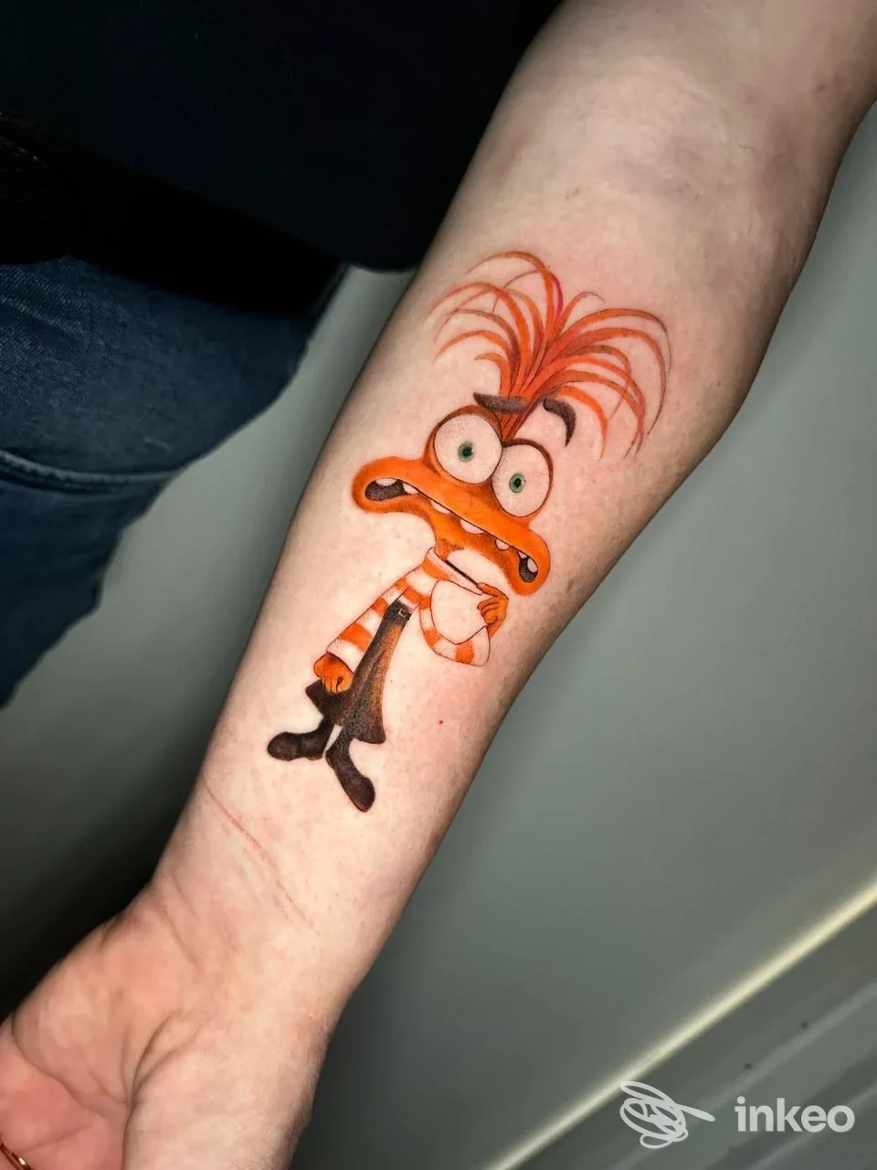 Tatuaggio su avambraccio raffigurante una mascotte cartoonesca con capelli arancioni e occhi grandi, in stile fumetto, eseguito sulla pelle reale, visualizzato in una foto singola.