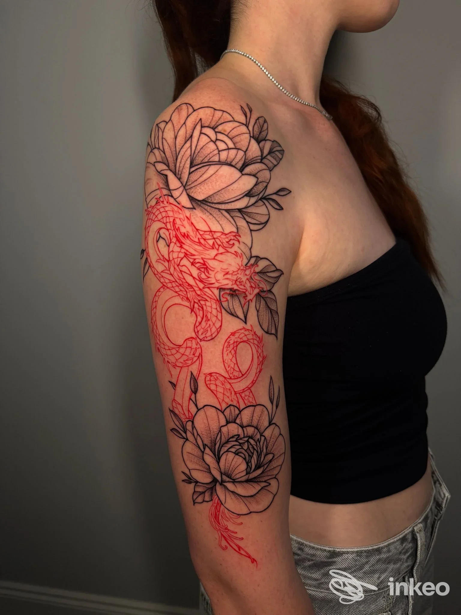 Spalla e braccio tatuati con grandi fiori neri e bozzetto di drago rosso; lavoro fotografato da Black Blood tattoo, Anguillara Sabazia.