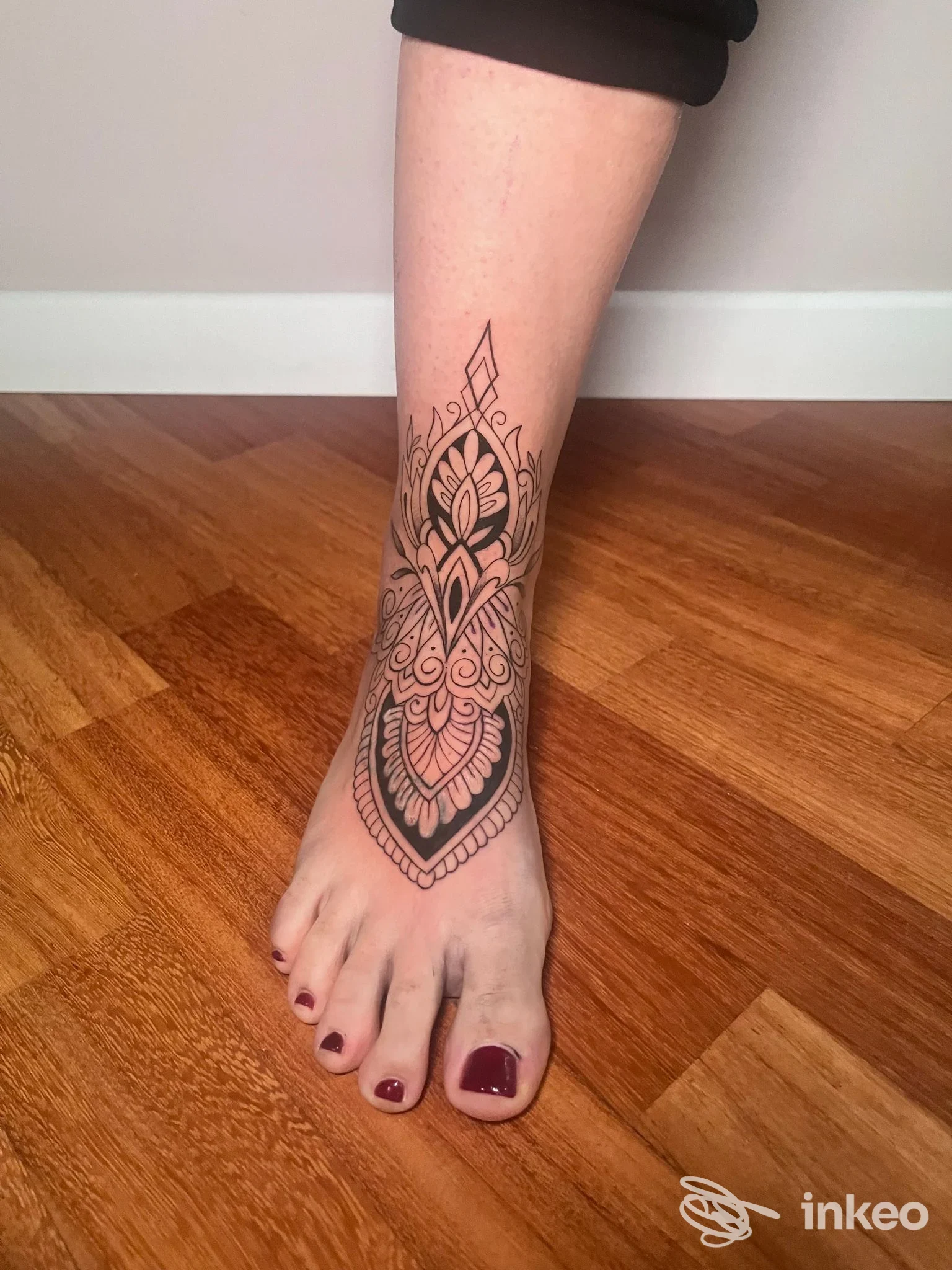 Tattoo in stile mandala lungo la caviglia e sul dorso del piede, realizzato su pelle; senza volto visibile, pavimento in legno sullo sfondo, un’unica inquadratura.