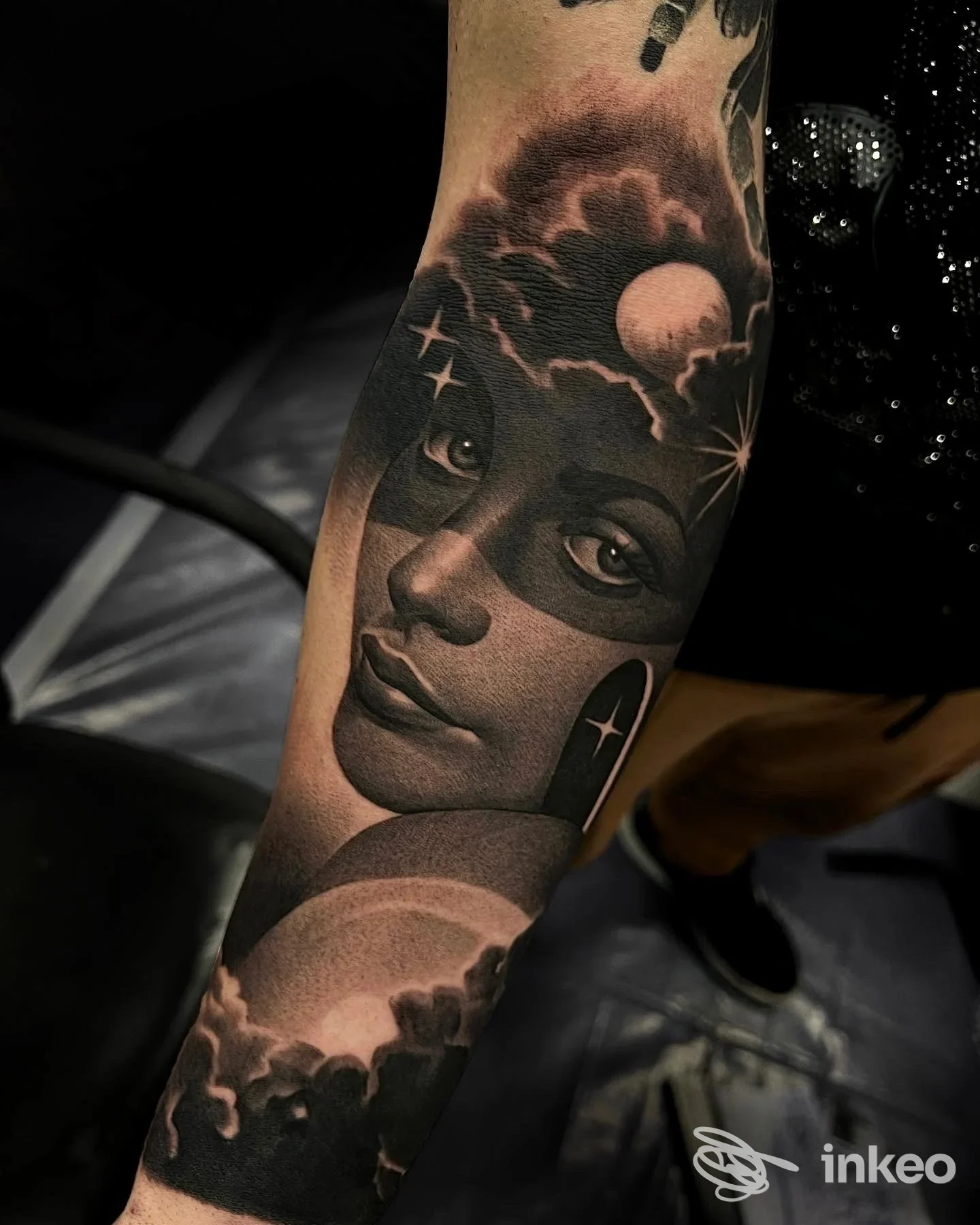 Tatuaggio realistico in bianco e nero di un volto surreale su avambraccio, con lune e nuvole; scatto professionale — Cromo Tattoo Studio, Roma.