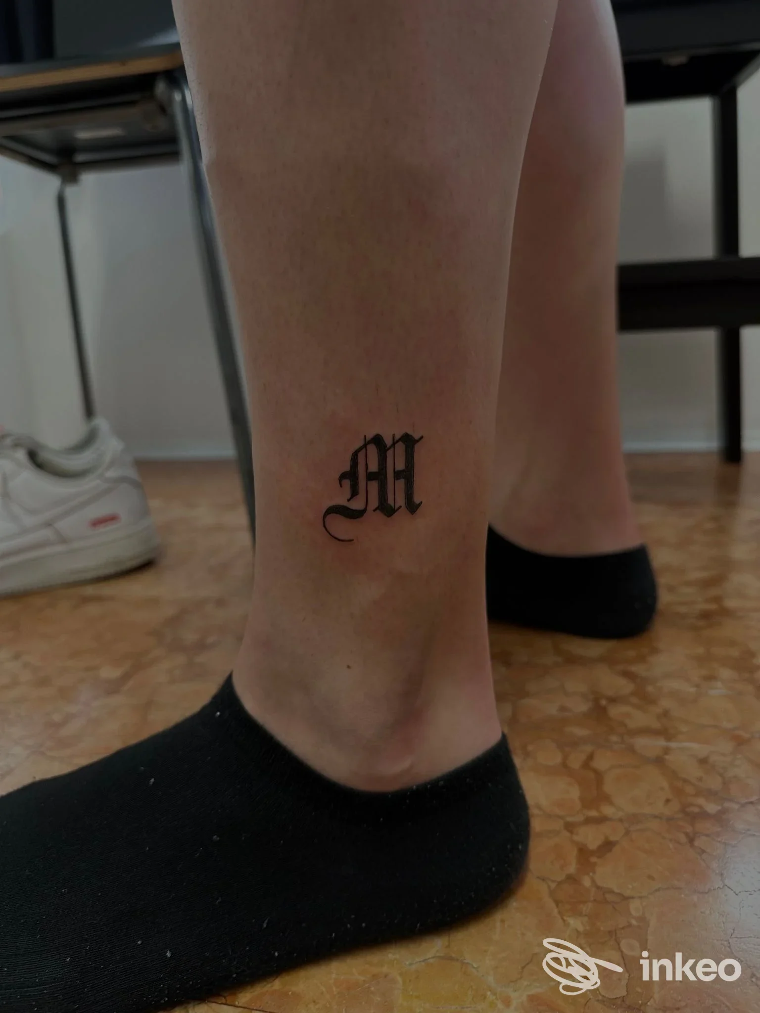 Tatuaggio in stile lettering gotico della lettera 'M' in nero posizionato sulla caviglia di una persona, Forma Mentis Tattoo Art, Sezze.