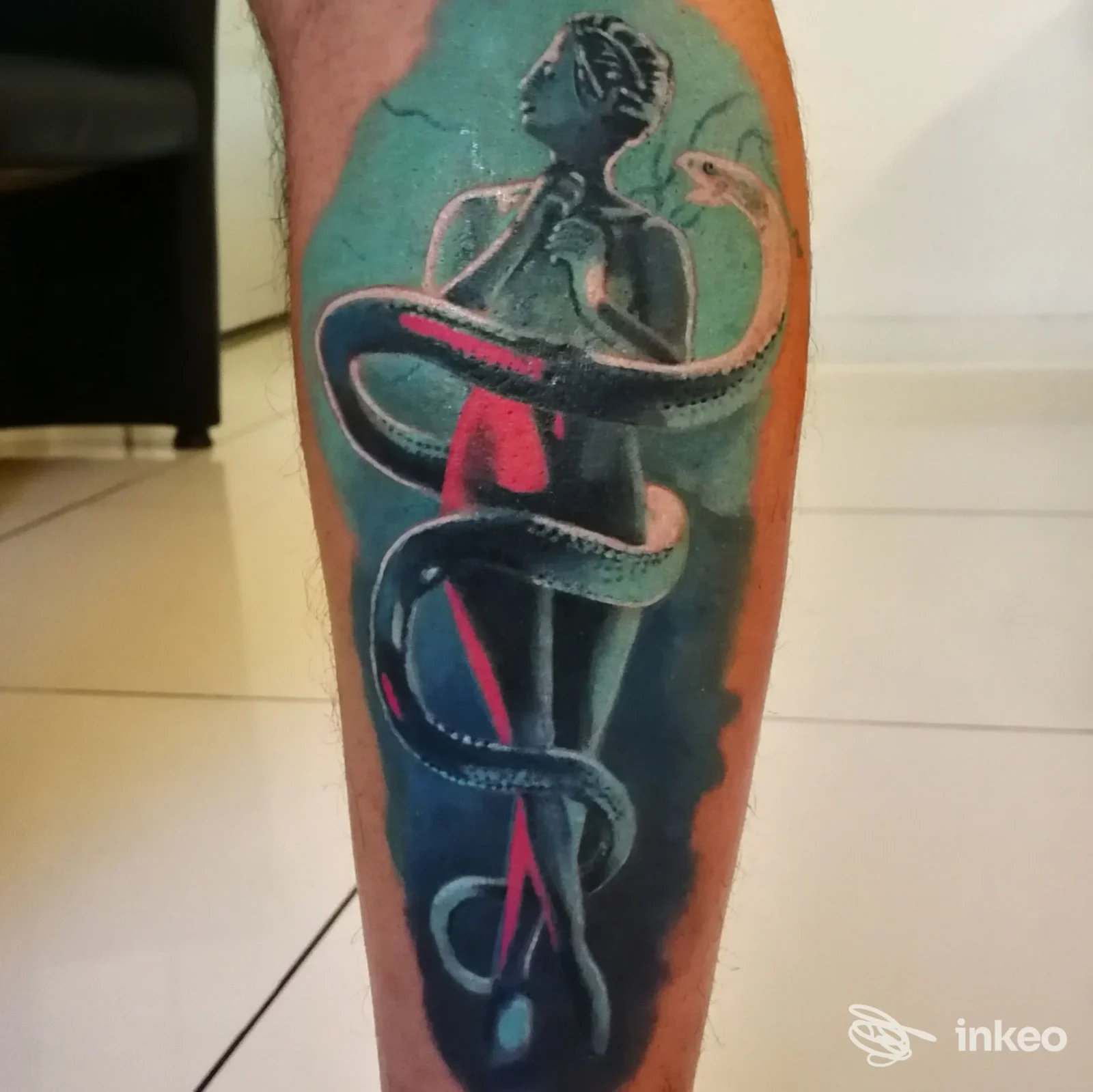 Tattoo a colori su pelle della gamba che mostra una figura umana stilizzata avvolta da un serpente, con sfondo azzurro-teal e accenti rosa, senza testo o watermark visibili.