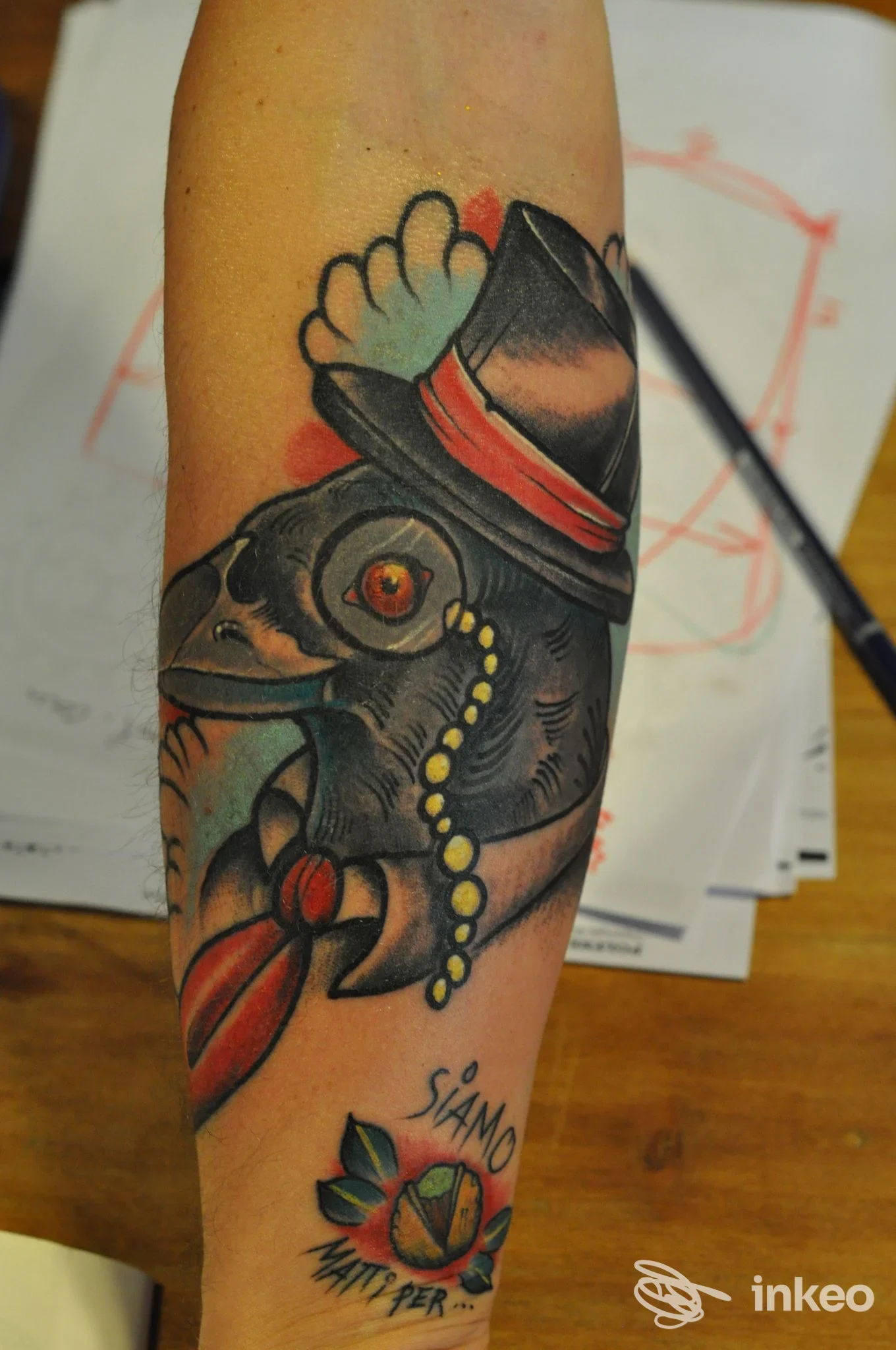 Tattoo su avambraccio raffigurante una creatura con cappello e collana, con colori scuri e accenti rossi.