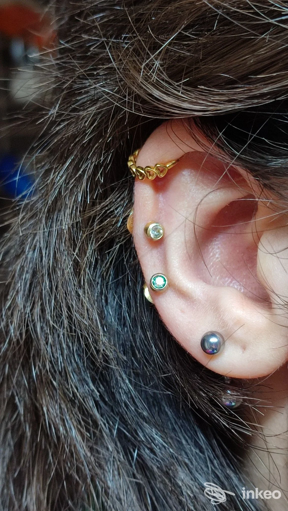 Piercing multipli sull'orecchio: catena dorata sull'helix e tre piercing con gemme lungo il lobo, scatto in primo piano su capelli scuri.