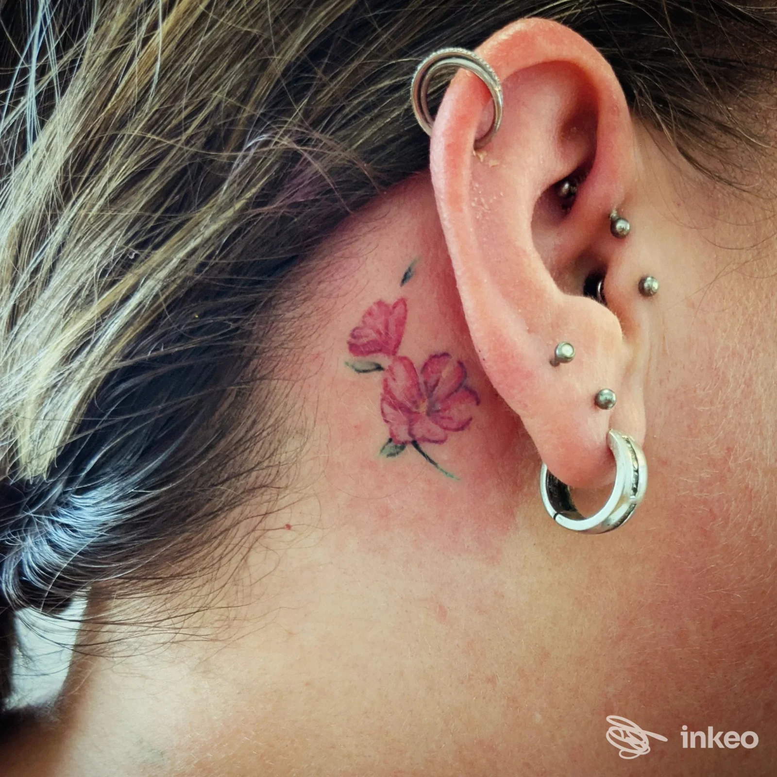 Piccolo tatuaggio floreale rosa dietro l'orecchio con piercings multipli, Alles Tattoo & Piercing Studio, Roma.