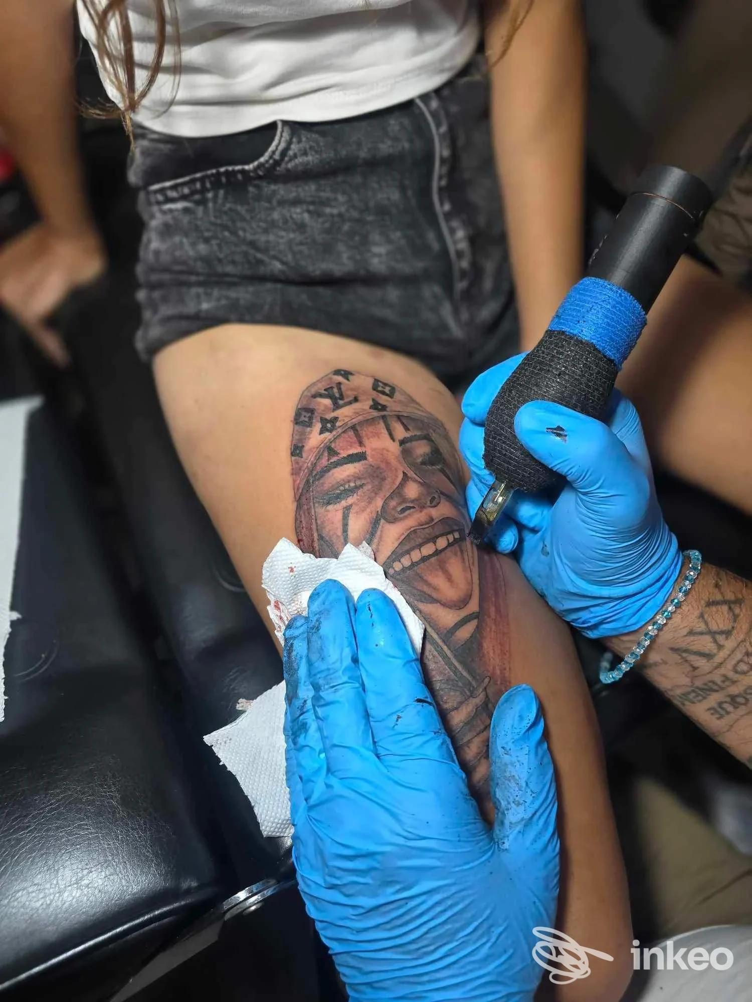 Tatuaggio realistico di un volto sulla coscia in corso di esecuzione, mani guantate del tatuatore visibili. Bottatattoo, Roma.