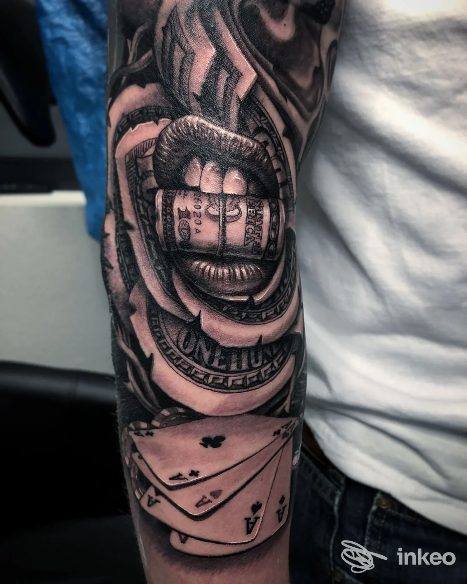 Tatuaggio realistico in bianco e nero su avambraccio con bocca che tiene una banconota e carte; Camdentown Tattoo Studio, Tivoli.