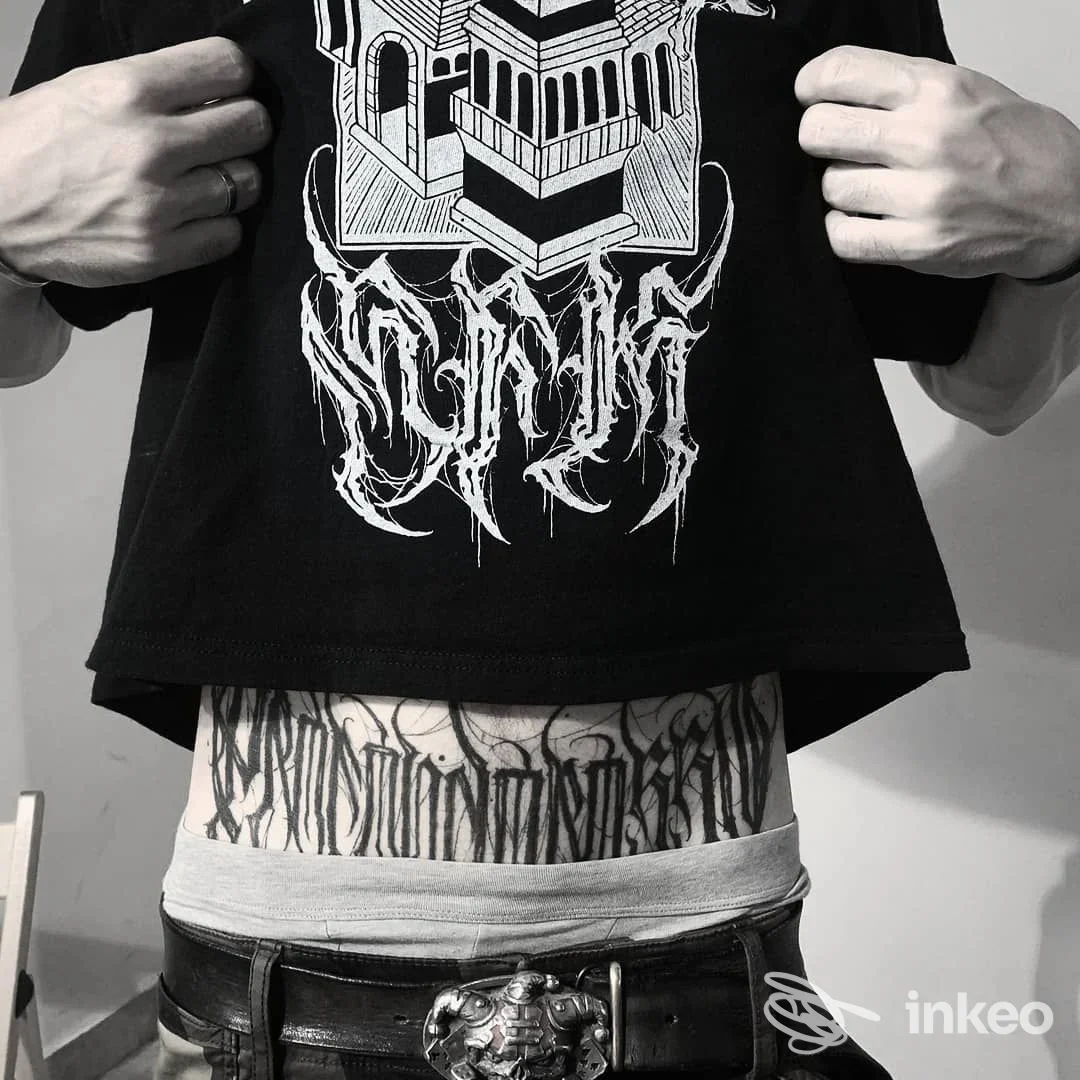 Dettaglio di tatuaggio blackwork/lettering sull'addome inferiore, scatto frontale che mostra il lavoro completo; Dinastia Fellas, Roma.
