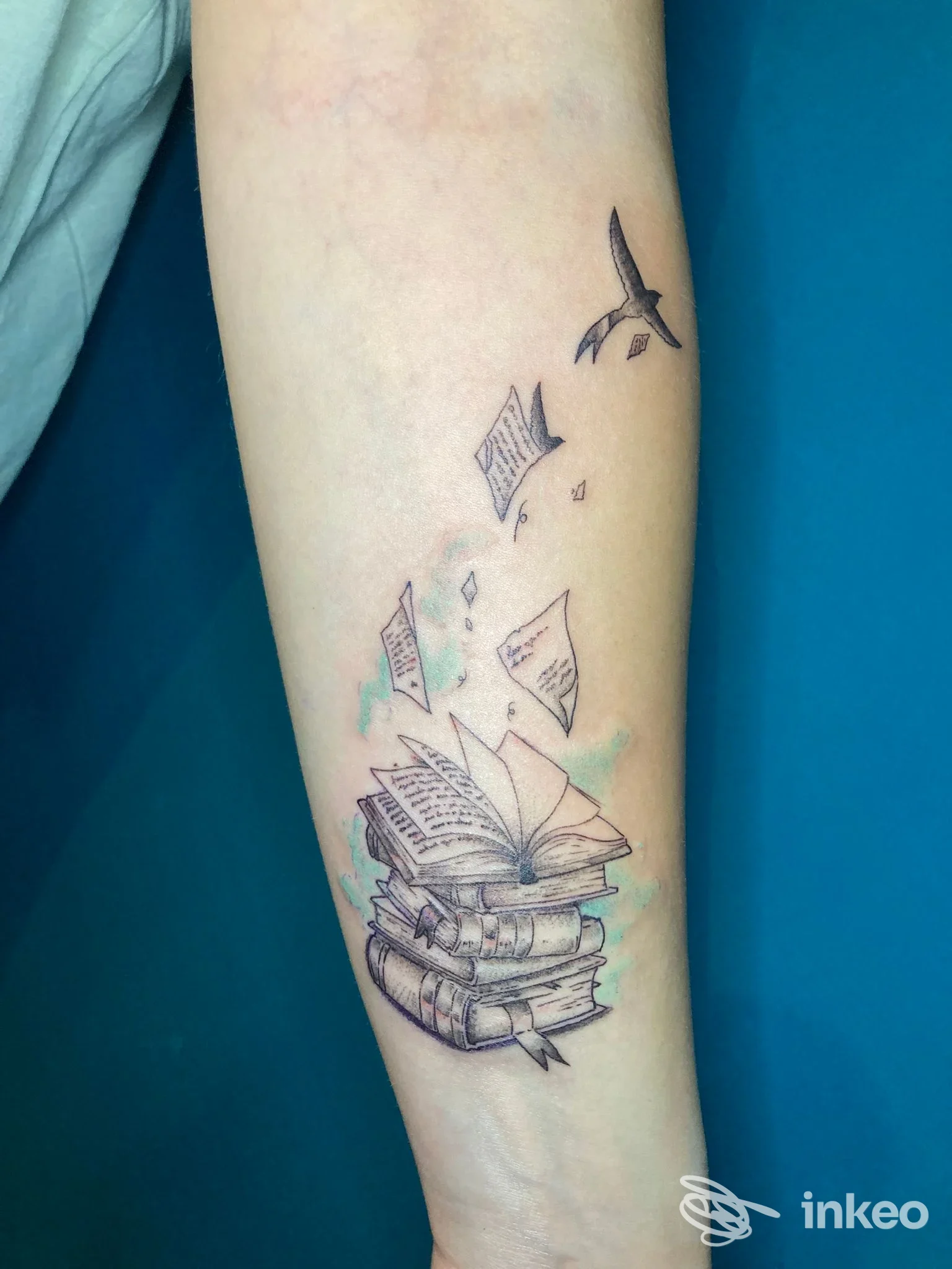 Tatuaggio su avambraccio con pila di libri, pagine volanti e un uccello, foto dallo studio Jelly Parson a Genzano di Roma.