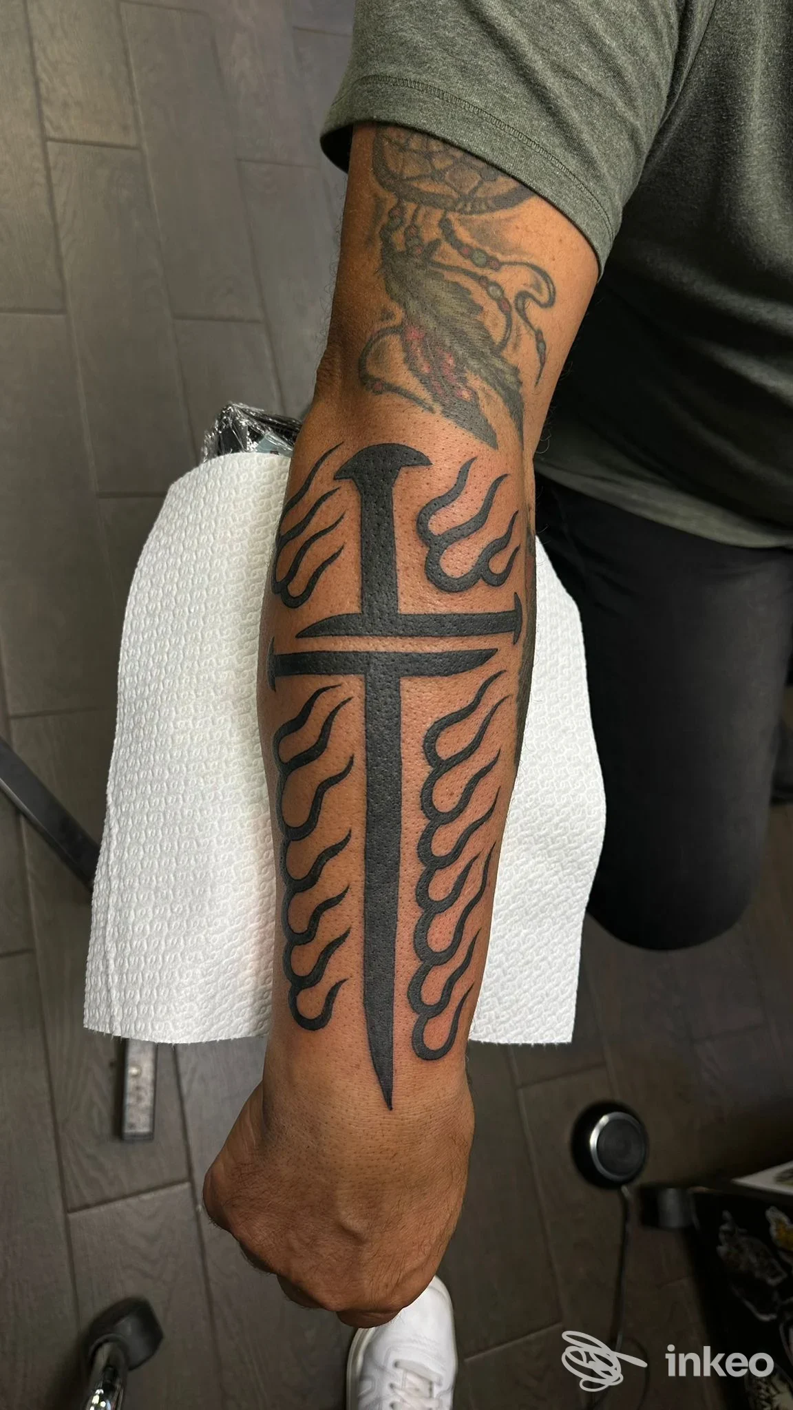 Avambraccio tatuato con una croce nera allungata e fiamme laterali, foto fornita da Traditional Stone Tattoo, Roma.