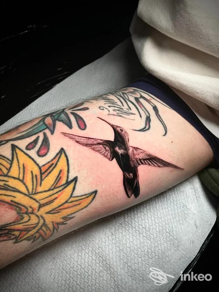 Tatuaggio di un colibrì nero e rosa sull'avambraccio, accanto a un grande fiore colorato. Foto fornita da TrameNoireInk Roma.