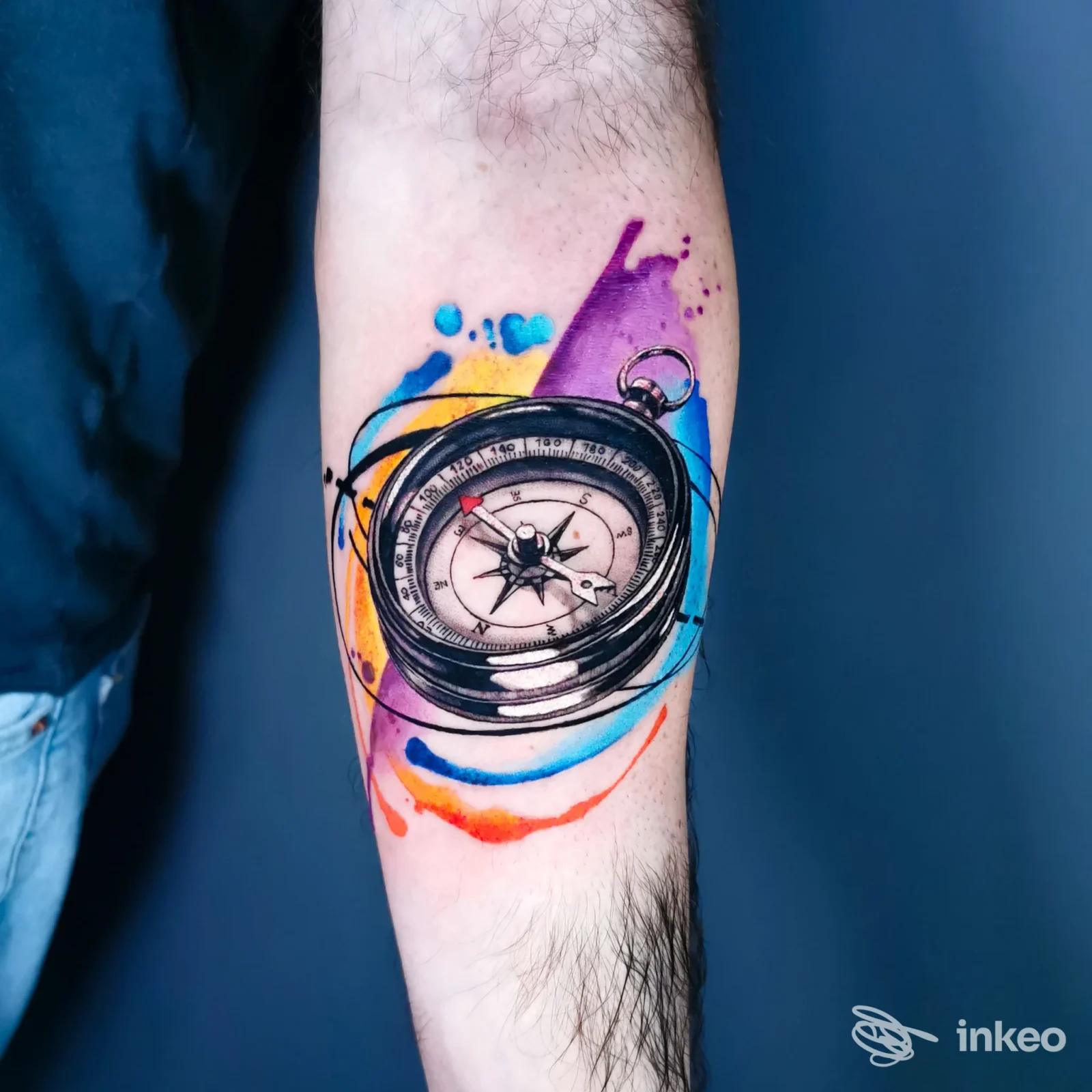 Avambraccio sinistro con tatuaggio a colori di una bussola, circondato da schizzi viola, blu, giallo e arancione, su pelle reale senza visione di volti.