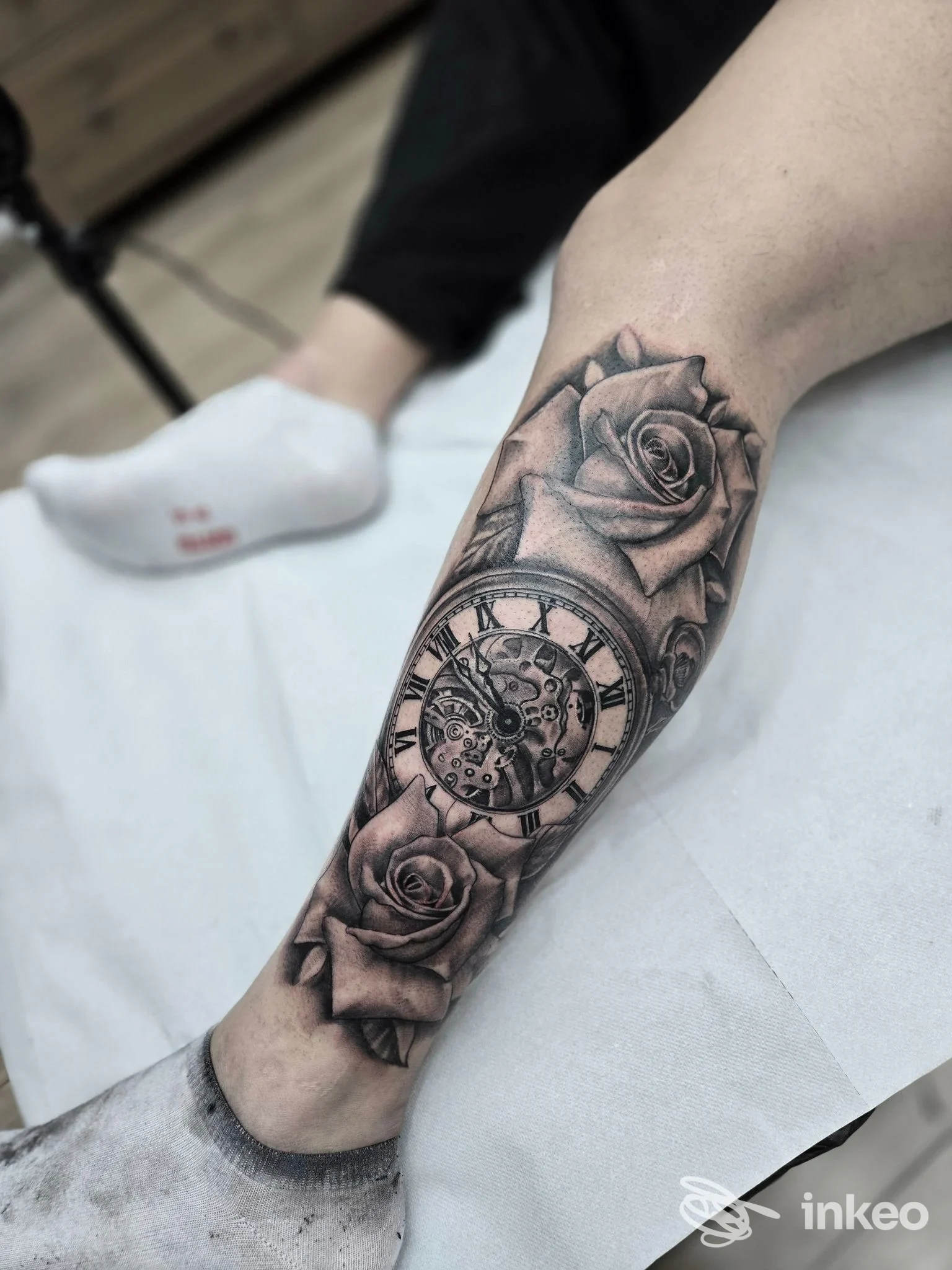 Tattoo sull’avambraccio sinistro, raffigurante un orologio rotondo con numeri romani, circondato da due rose, in bianco e nero, visibile sulla gamba del modello.
