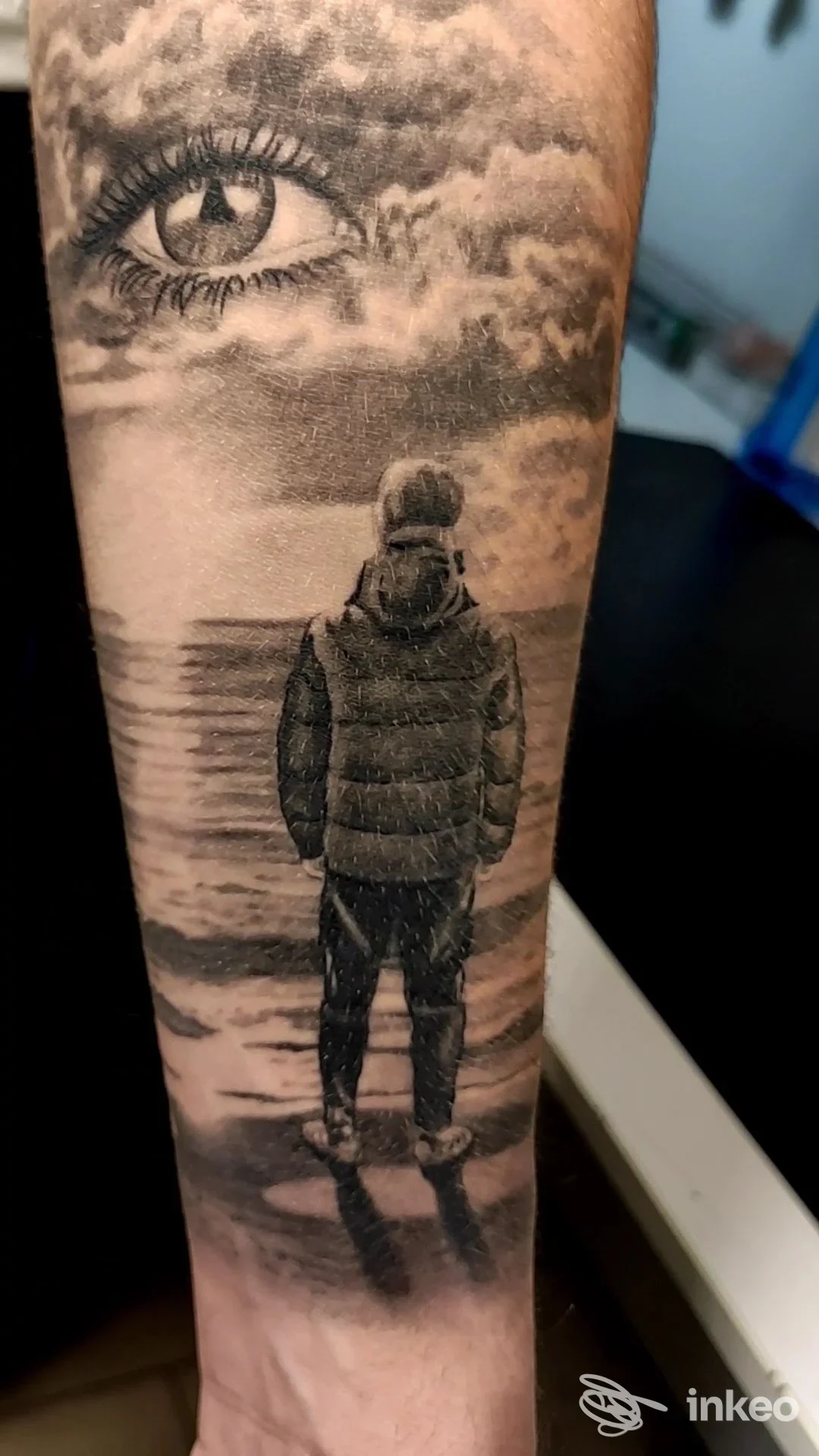 Tatuaggio realistico in bianco e nero su avambraccio: grande occhio tra le nuvole e figura di spalle verso il mare. Derek Smith Tattoo studio, Pietra Ligure.