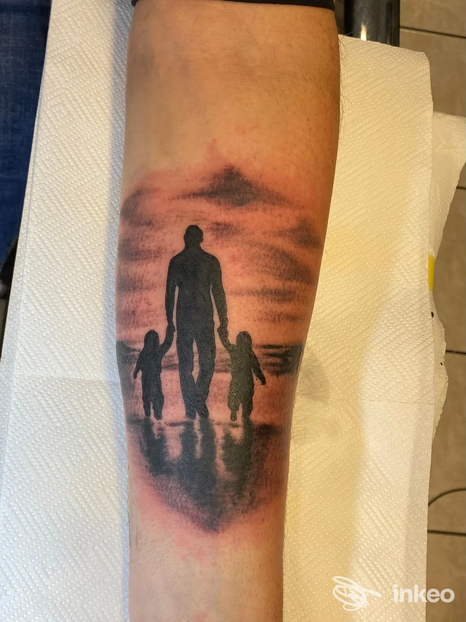 Tatuaggio sull'avambraccio di J Tattoo La Spezia (La Spezia): silhouette di un adulto che tiene per mano due bambini su riflessi da tramonto.