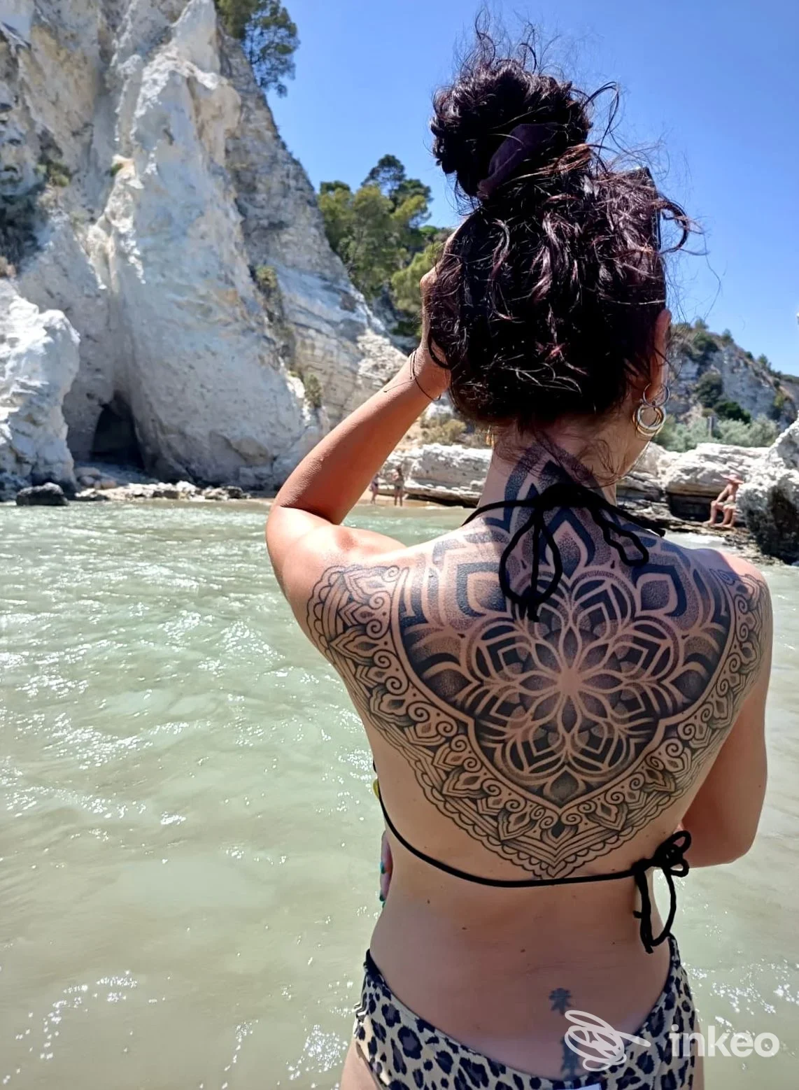 Persona vista di schiena mostra un grande tatuaggio nero a mandala sulla schiena alta, tra scapole e linea della cintura, con motivi geometrici.; lo sfondo mostra una costa rocciosa e mare.