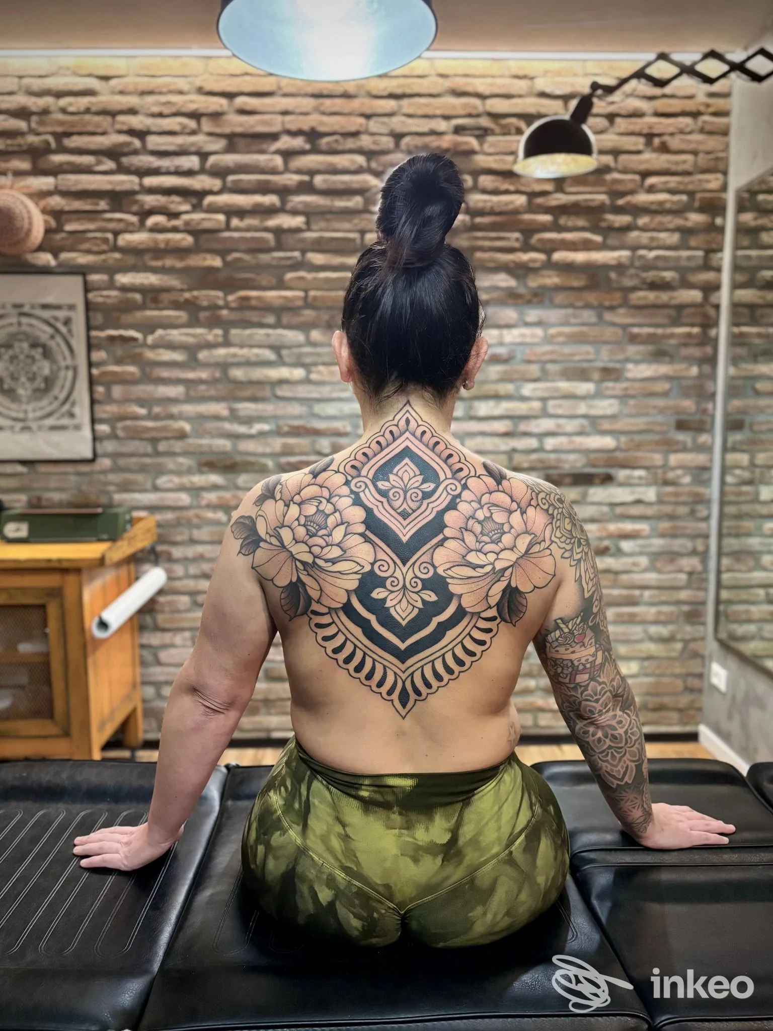Retro della schiena di una persona seduta con un grande tattoo geometrico floreale, visibile tra le scapole, in studio con pareti in pietra e illuminazione. s