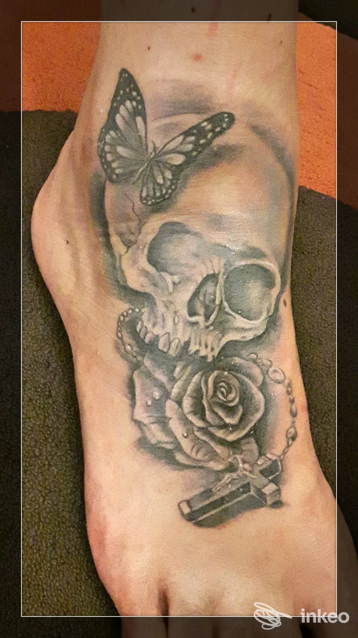 Primo piano del dorso del piede con tatuaggio realistico black & grey: teschio, rosa, farfalla e ciondolo. La Casa Del Tatuaggio, Luni