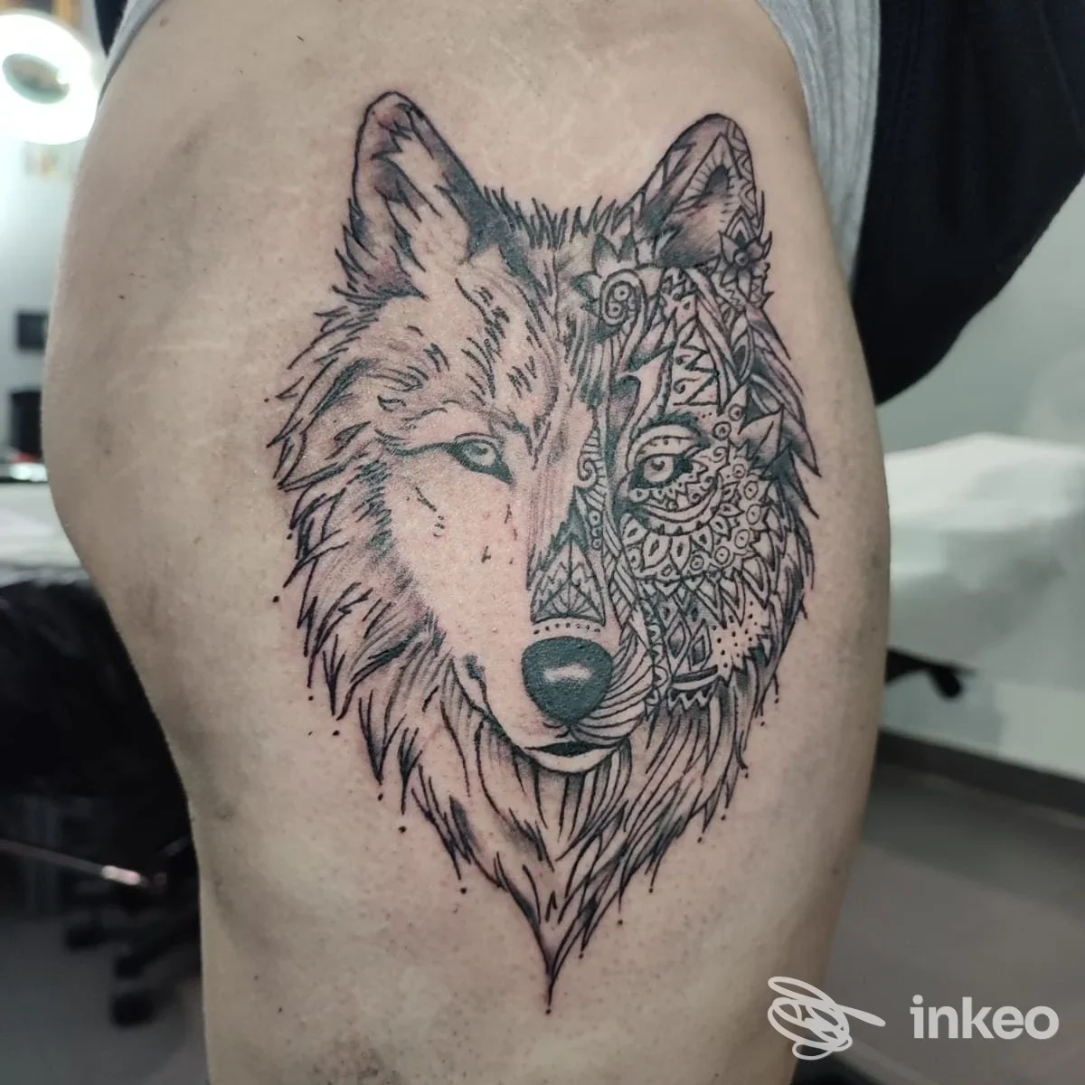 Tatuaggio grande a inchiostro nero di una testa di lupo, metà realistica e metà ornamentale, sulla coscia. Inkplosion Tattoo, Genova.