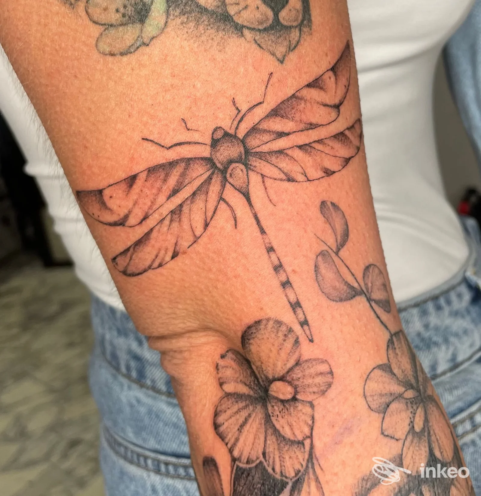 Tatuaggio a inchiostro nero con una libellula e fiori sull'avambraccio, foto fornita da Pink fluid tattoo studio ad Arenzano.