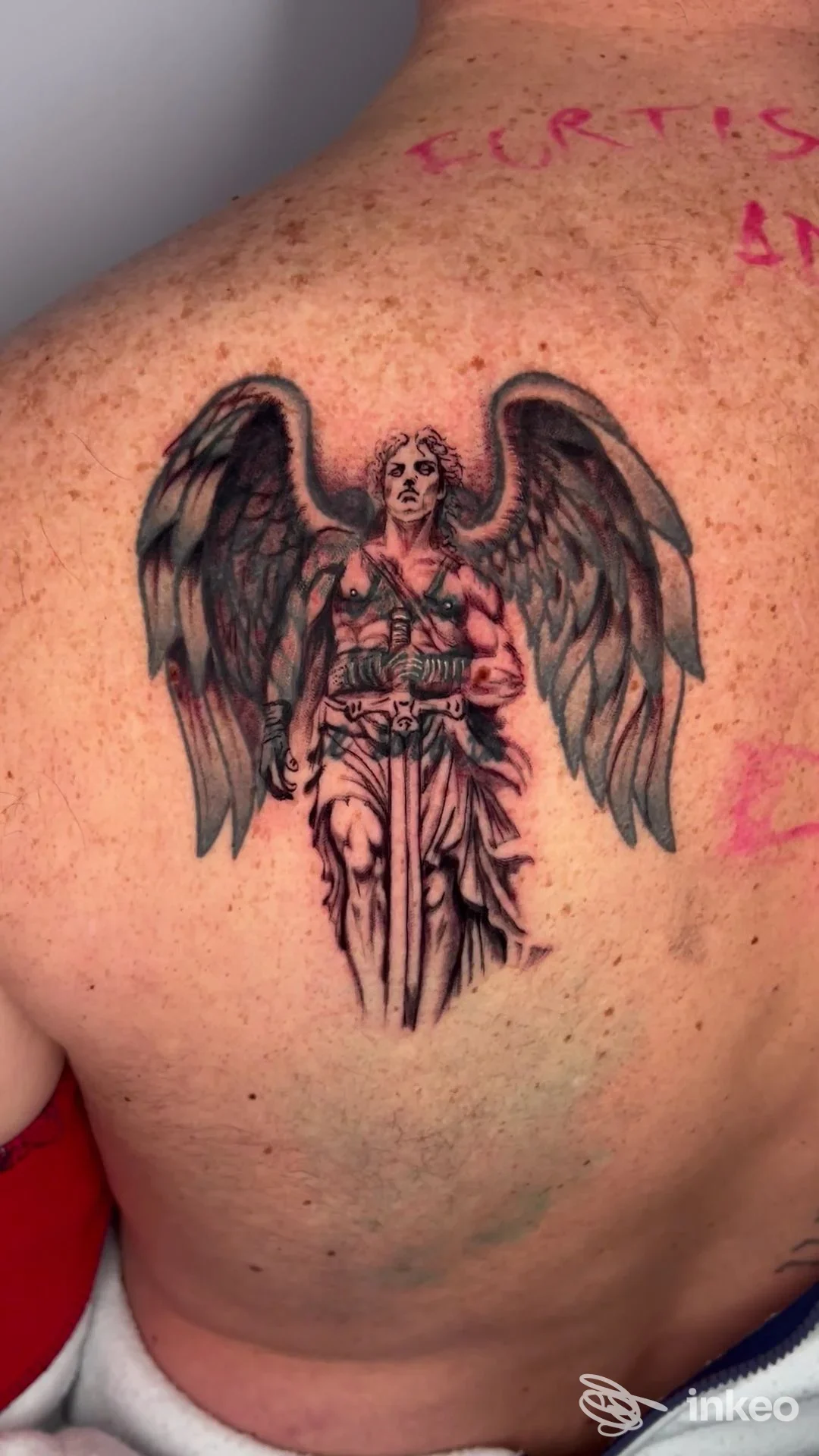 Tattoo realistico di un angelo con ali e spada sulla schiena, visibile in fotografia singola, senza overlay o testo digitale.