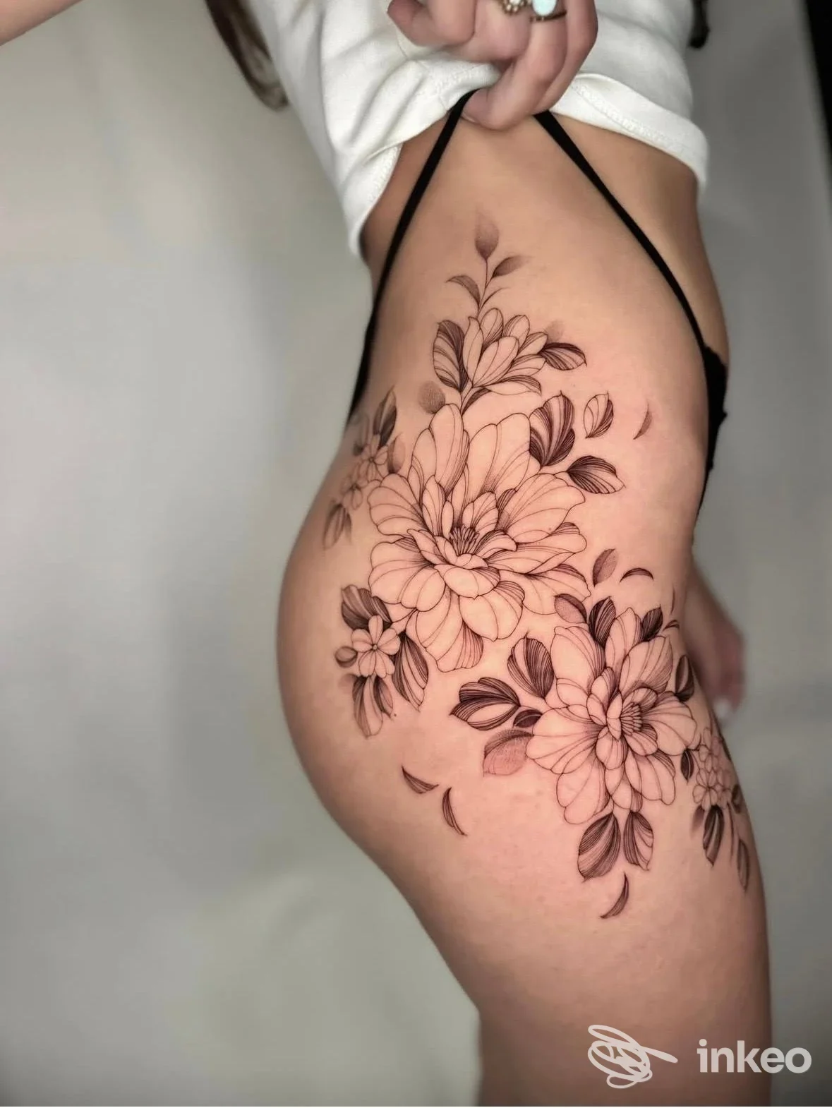 Tattoo floreale su coscia sinistra: fiori stilizzati in nero/grigio su pelle chiara, con dettagli di foglie; scatto laterale che mostra chiaramente il disegno completo sulla pelle.